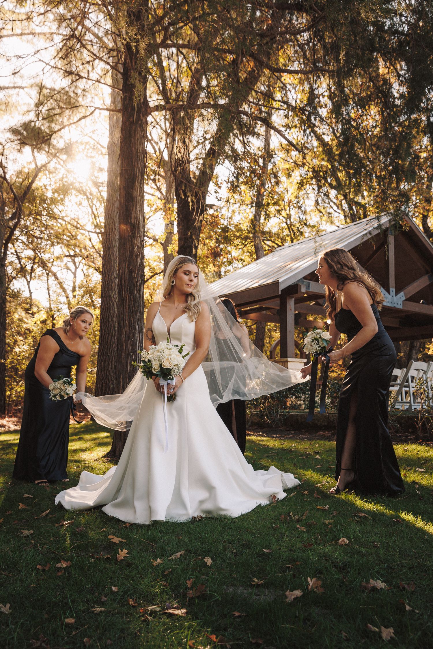 Bride in white gown with veil held by bridesmaids in black dresses at outdoor wedding.