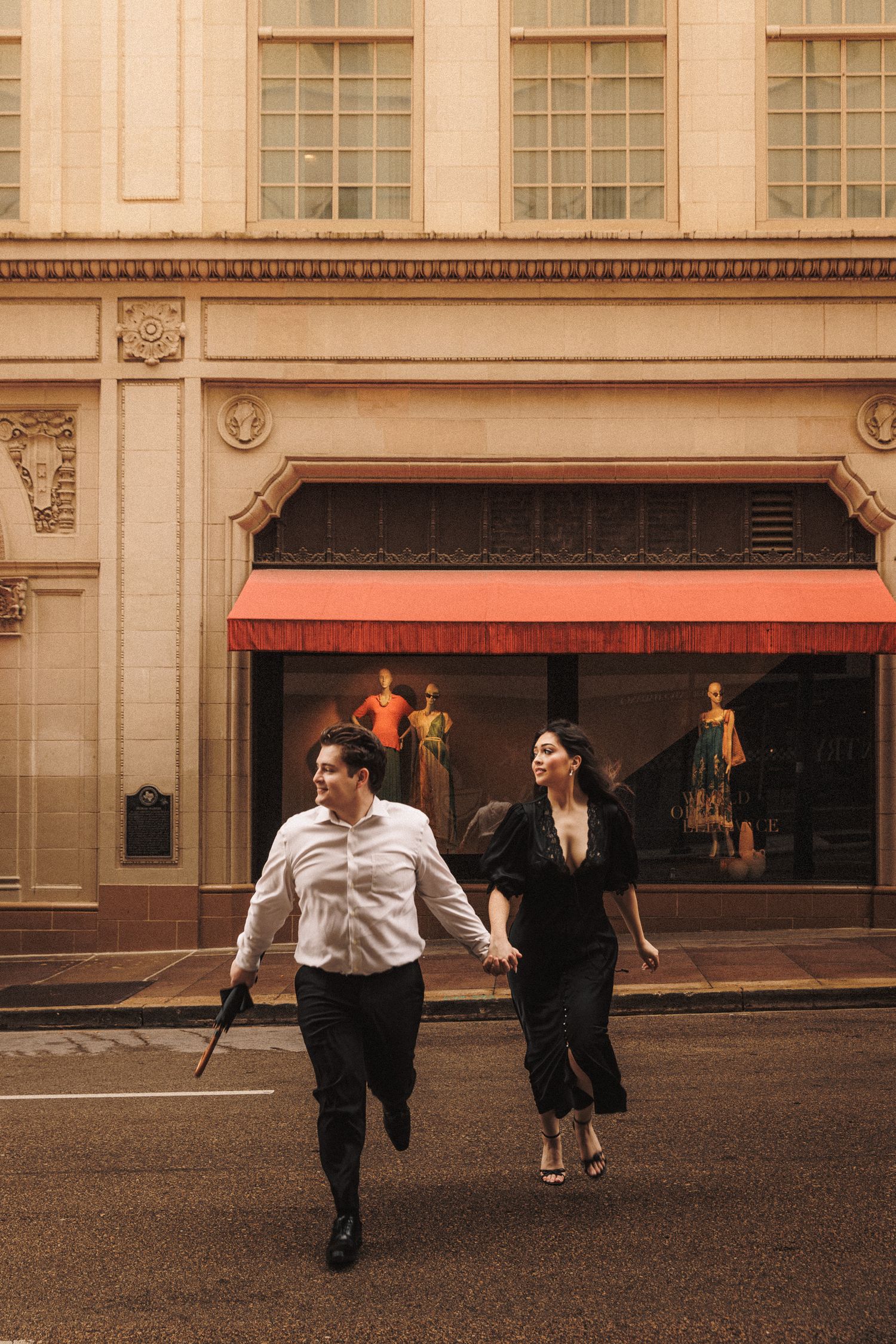 A stylish couple runs hand-in-hand across a city street in front of an elegant storefront.