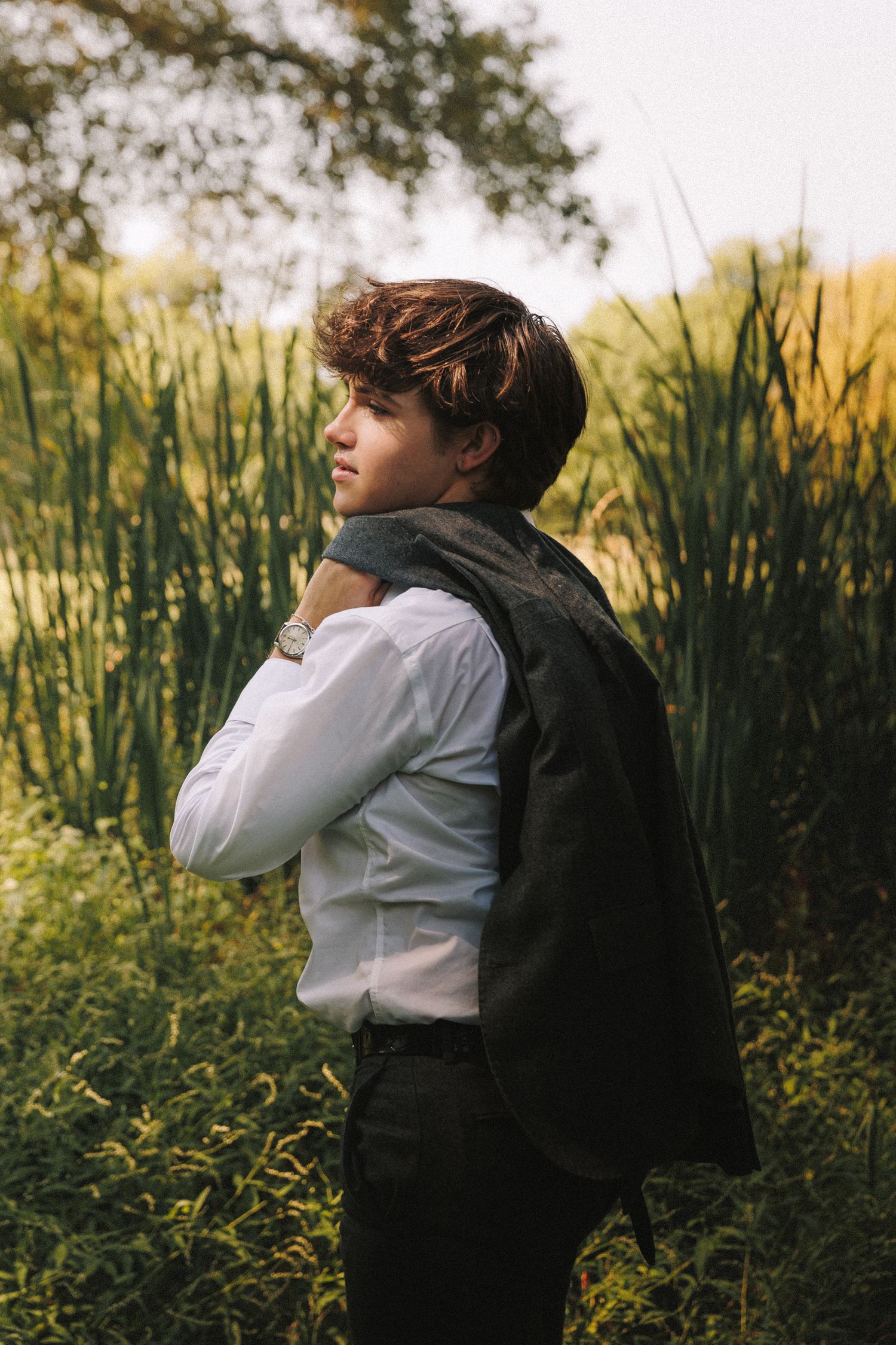 Person in white shirt and black jacket poses against natural backdrop of tall reeds and wildflowers at sunset.