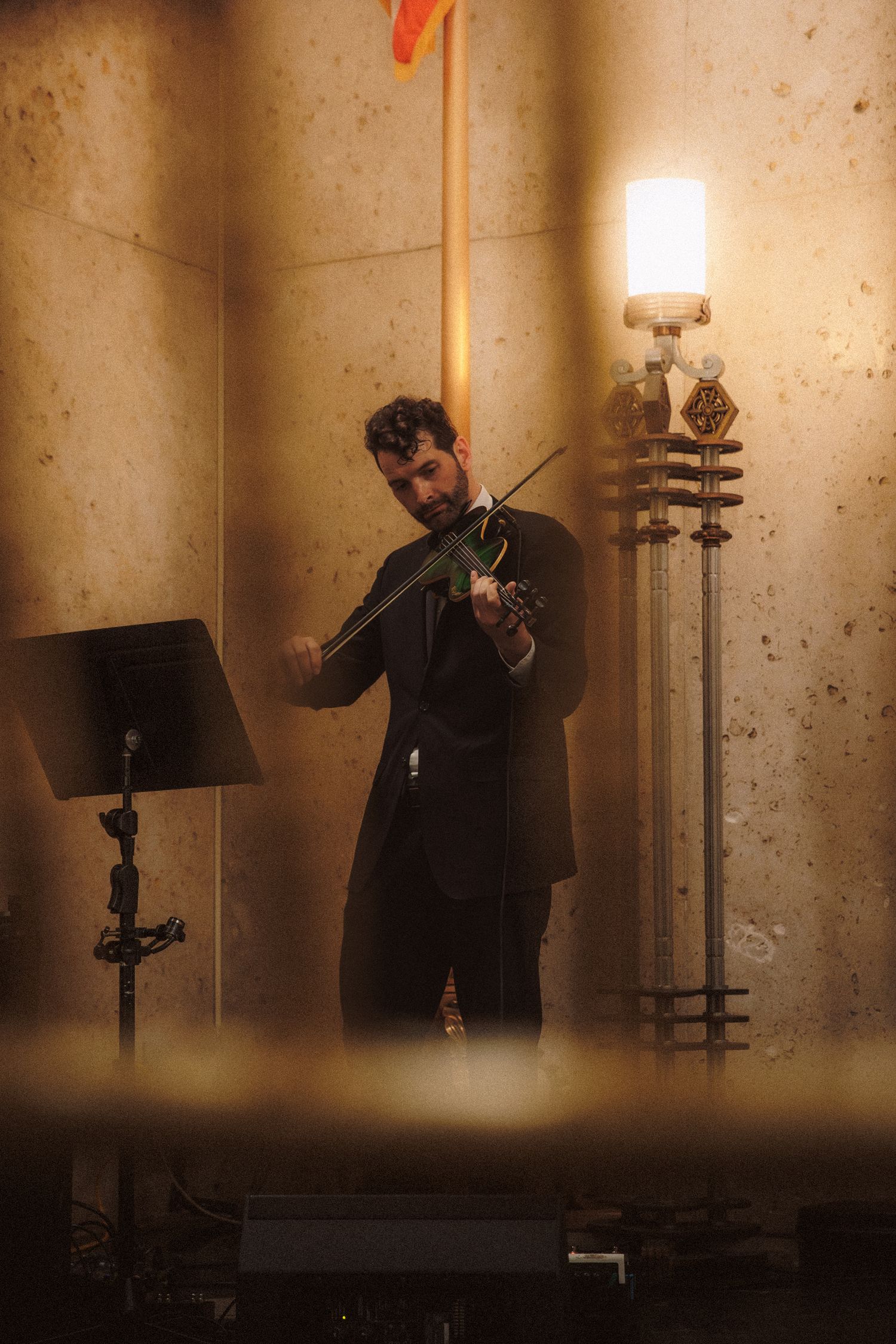 Male violinist in black suit performing near ornate wall sconce in elegant stone venue.