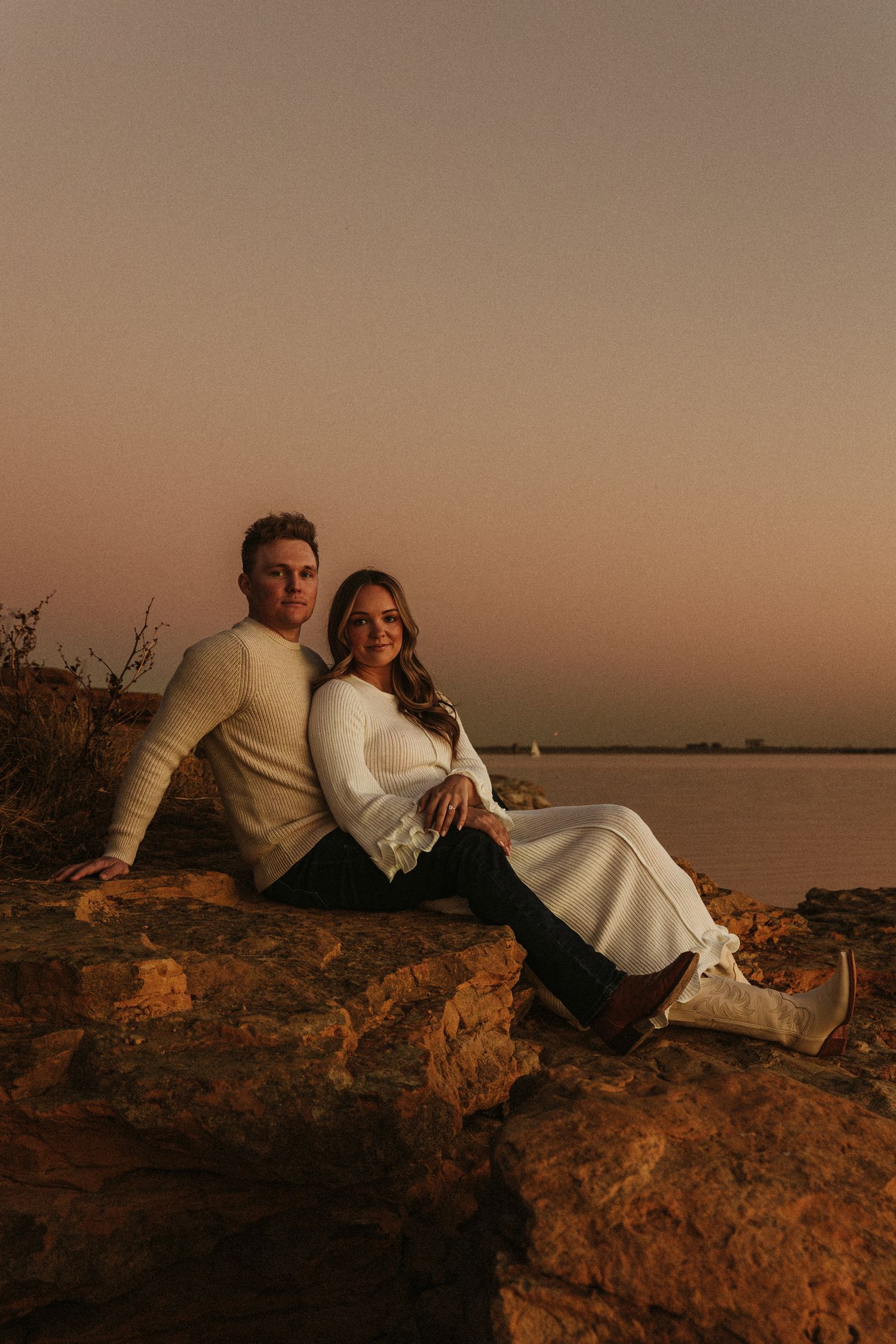 Couple relaxing on rocky cliff overlooking a tranquil lake at dusk.