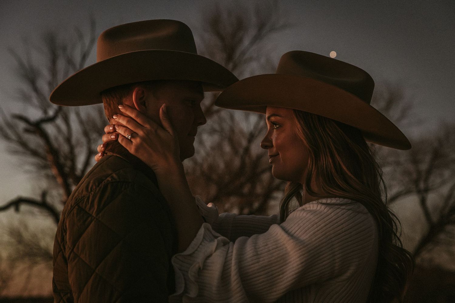Silhouetted figures in cowboy hats share an intimate moment against a dusky evening sky.