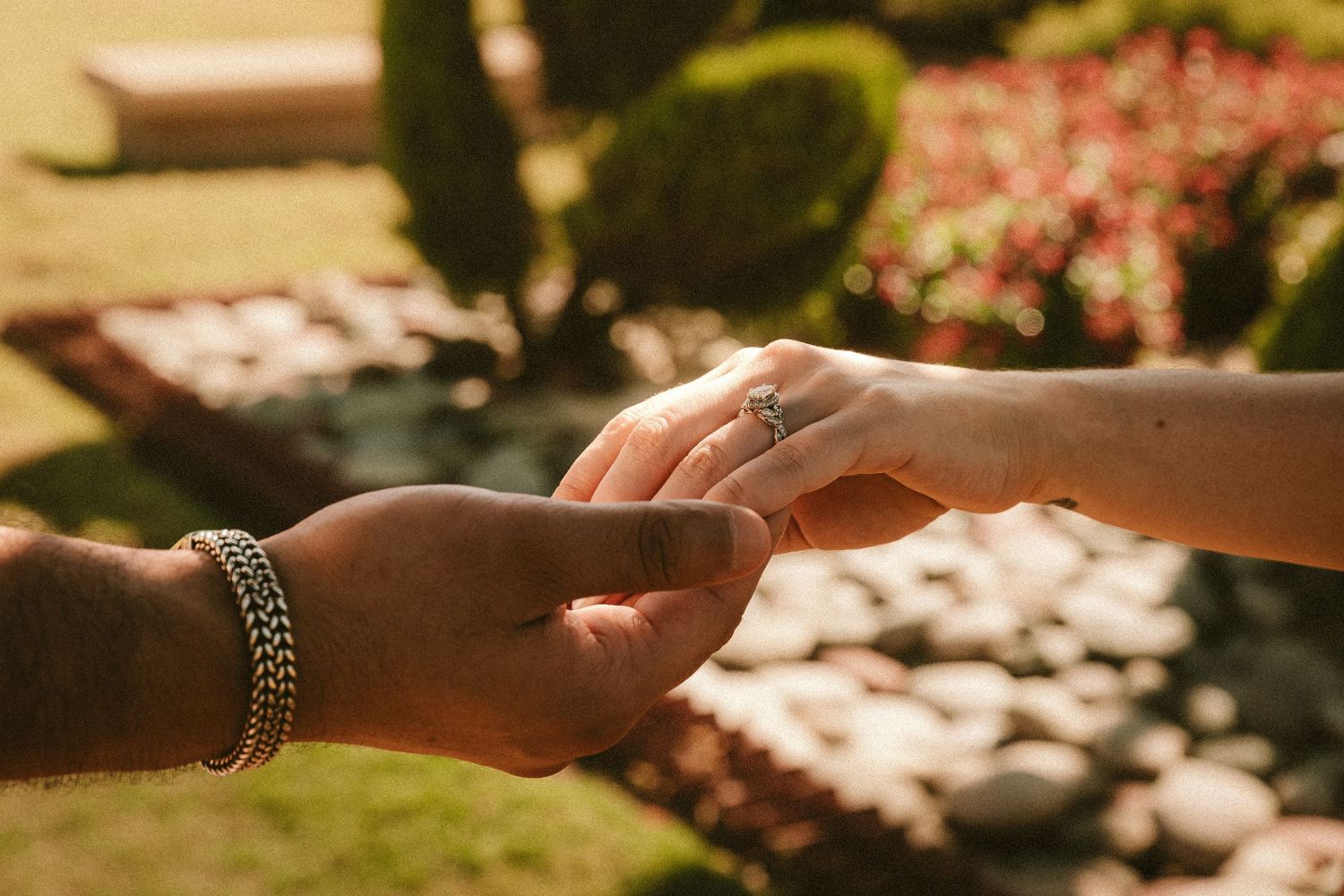 Close up view of hands exchanging wedding rings during outdoor ceremony with blurred floral background.
