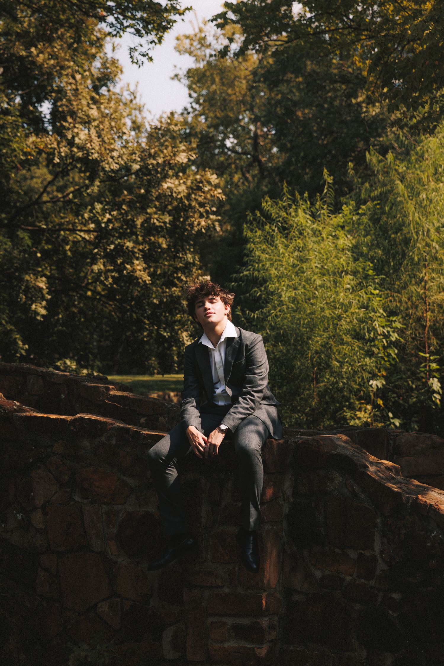 A person in a suit sits on a stone wall surrounded by autumn trees in a park setting.