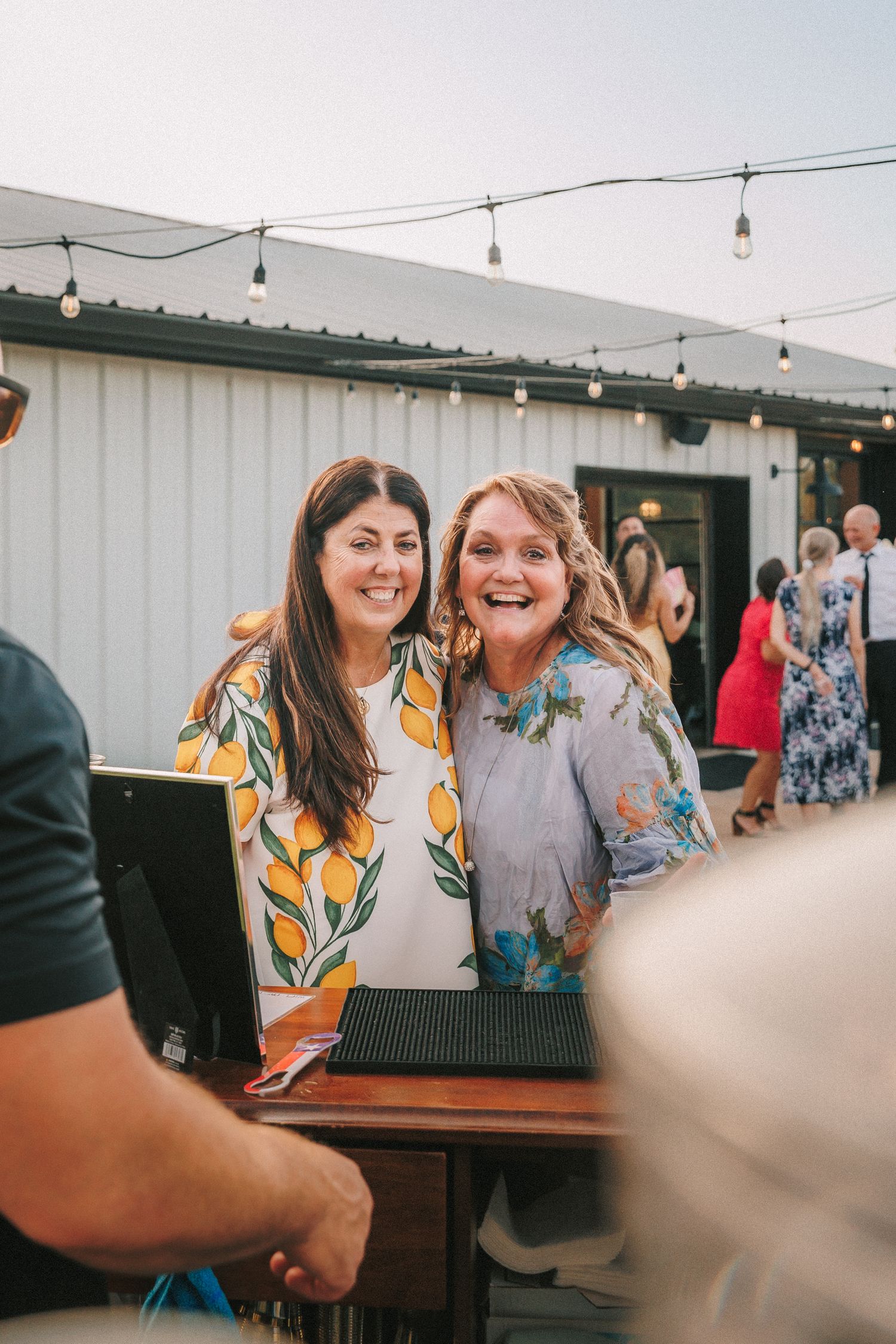 Wedding guests socializing and laughing at an outdoor venue with string lights overhead.