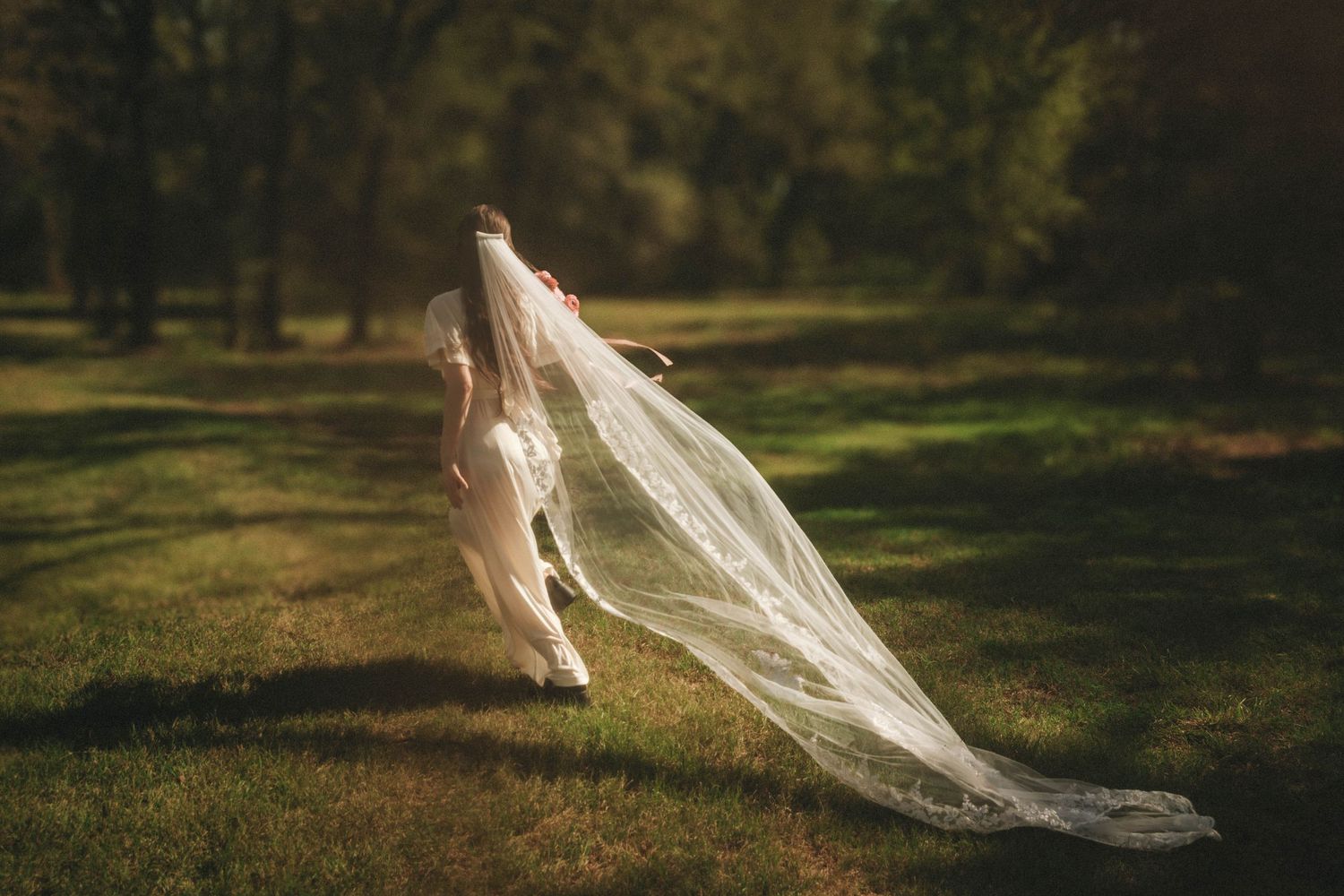 A bride in a flowing white wedding dress and long veil stands in a dreamy sunlit forest setting.