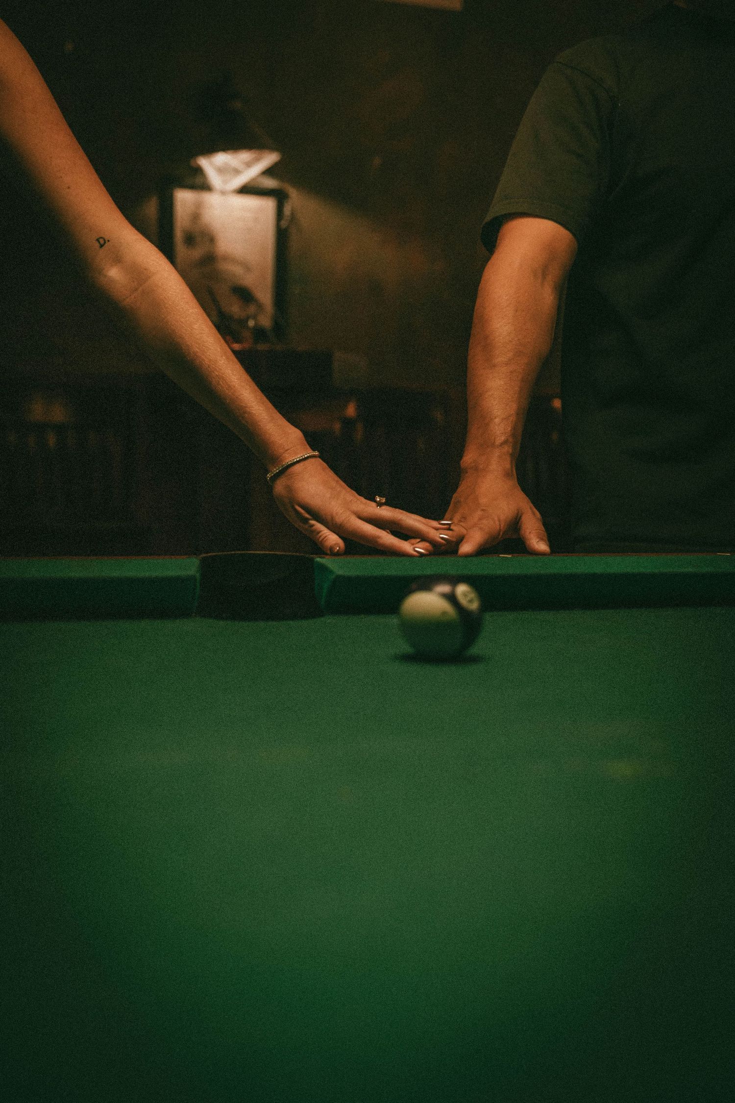 Close-up of hands playing pool on a green felt table in dim lighting.