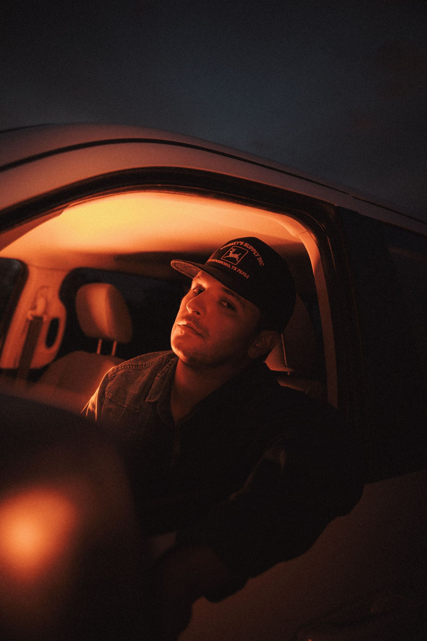 A person in a baseball cap sits in a car illuminated by warm orange light creating a moody nighttime atmosphere.
