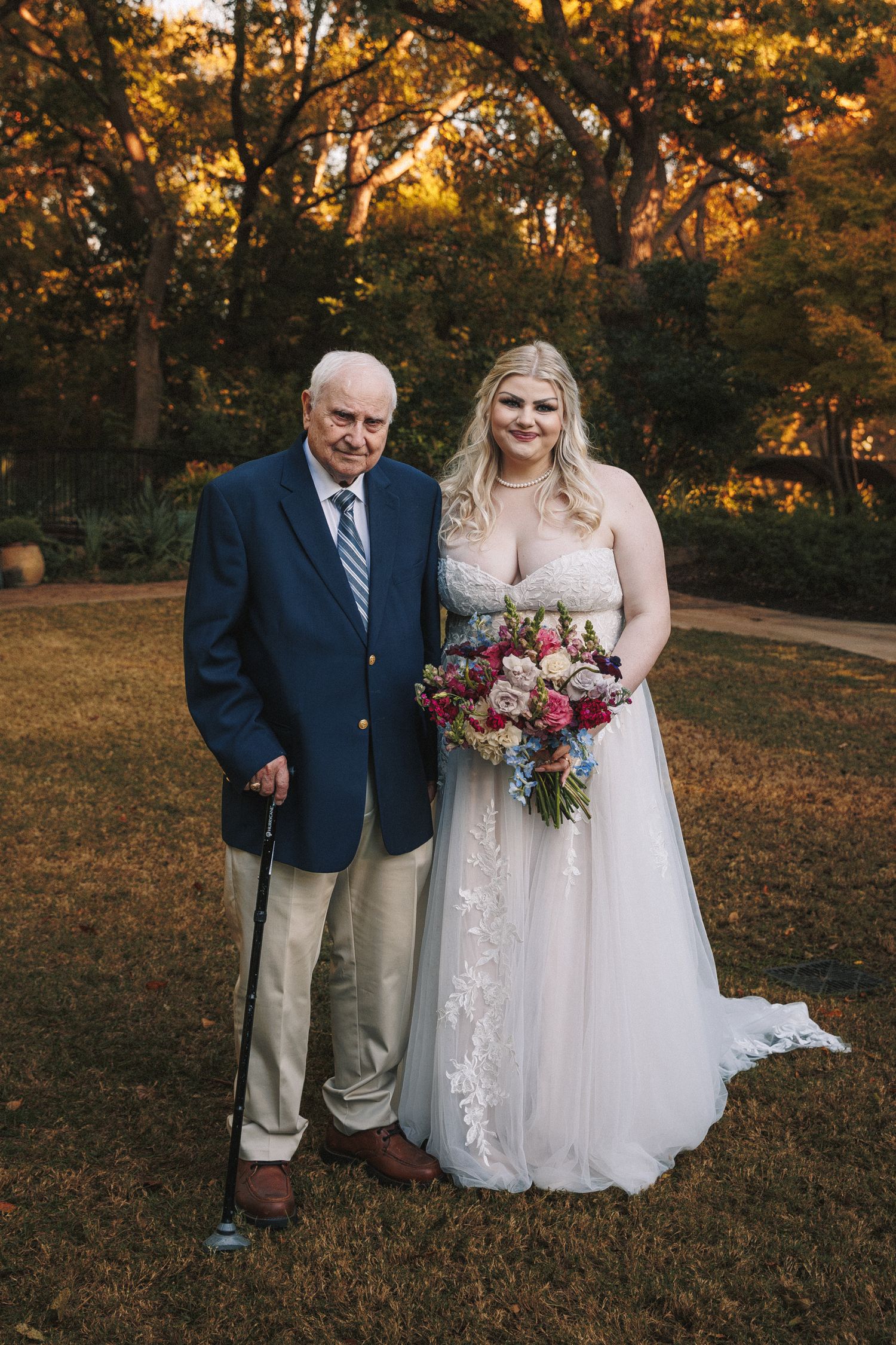 Bride in lace wedding gown holding colorful bouquet posing with elderly grandfather outdoors.
