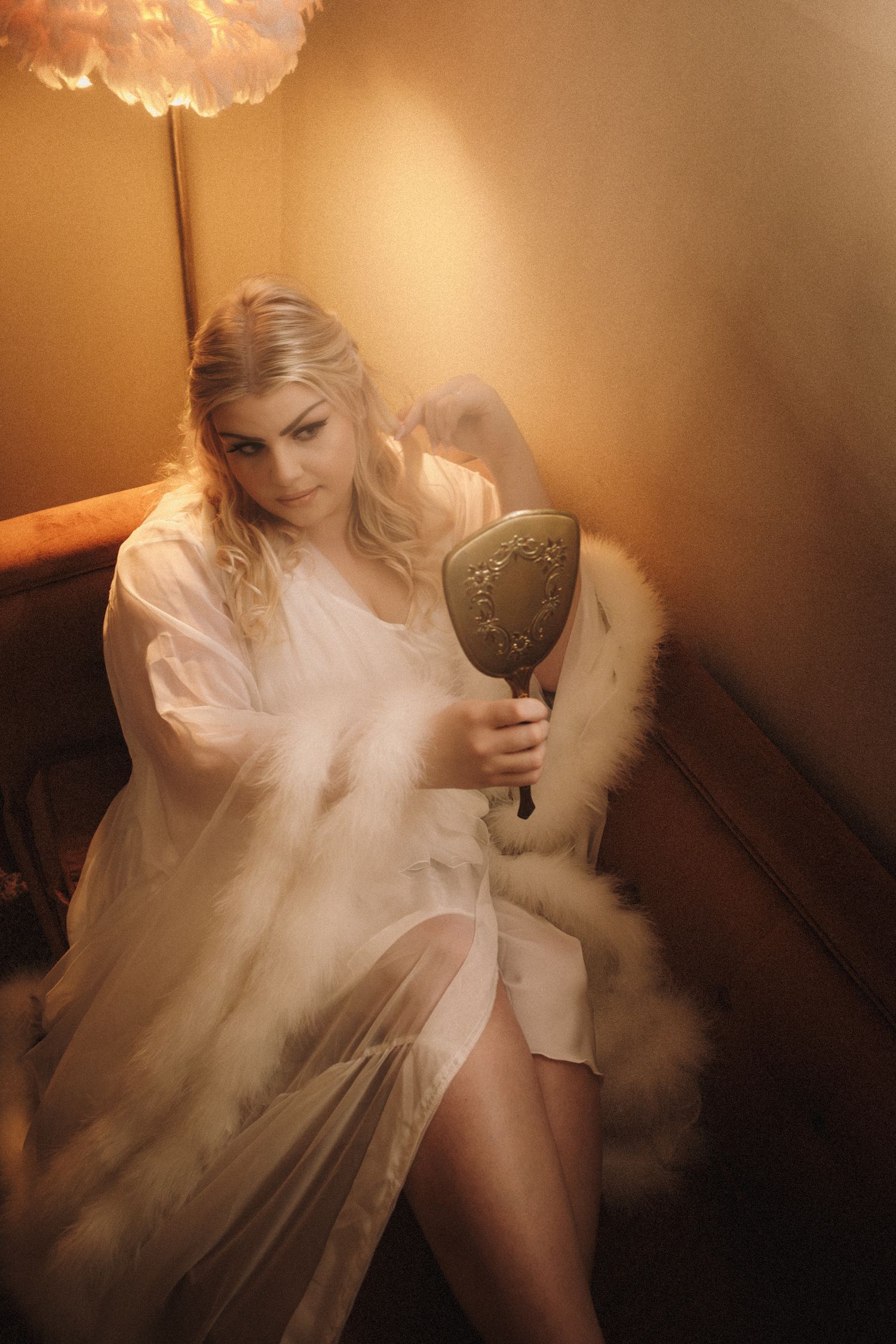 Blonde bride in white sheer robe holding vintage hand mirror while seated in warmly lit room.