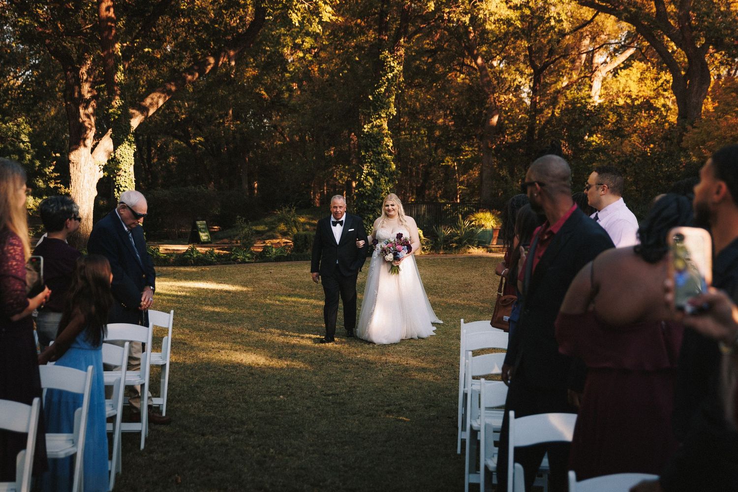Bride walks down outdoor aisle with escort at sunset wedding ceremony with white chairs.