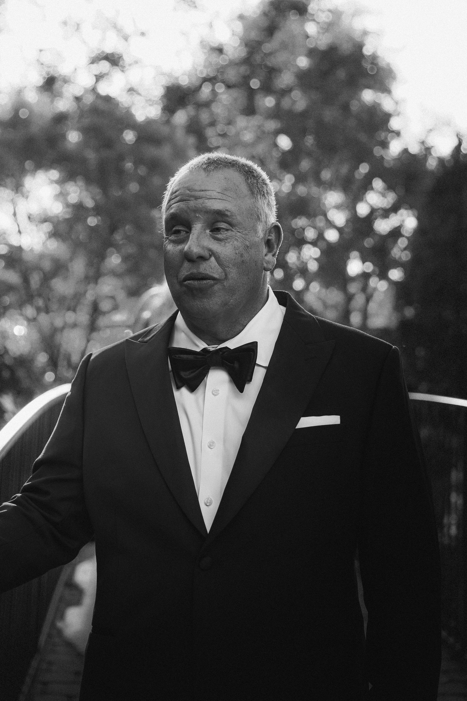 Distinguished man in black tuxedo and bow tie posing outdoors in black and white portrait.