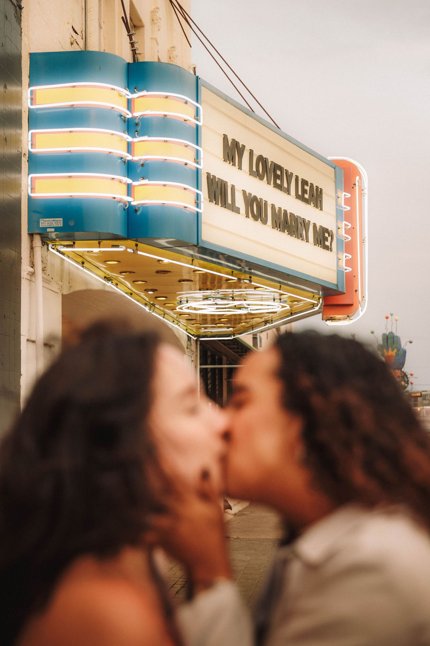 Couple shares a kiss in front of glowing marriage proposal marquee sign at a vintage theater.