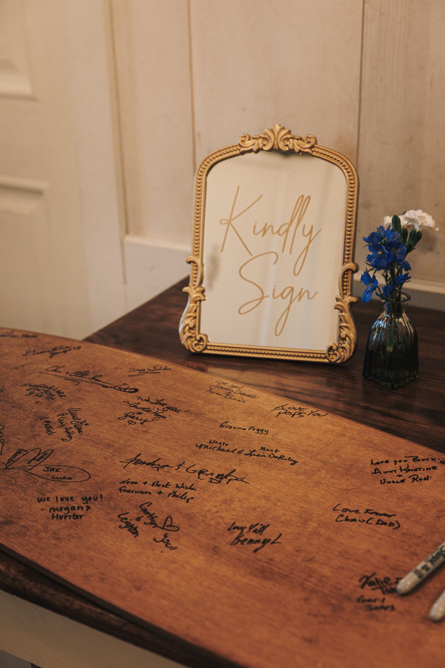 Wooden guest book signing table with a gold-framed Kindly Sign acrylic sign and blue flower vase.