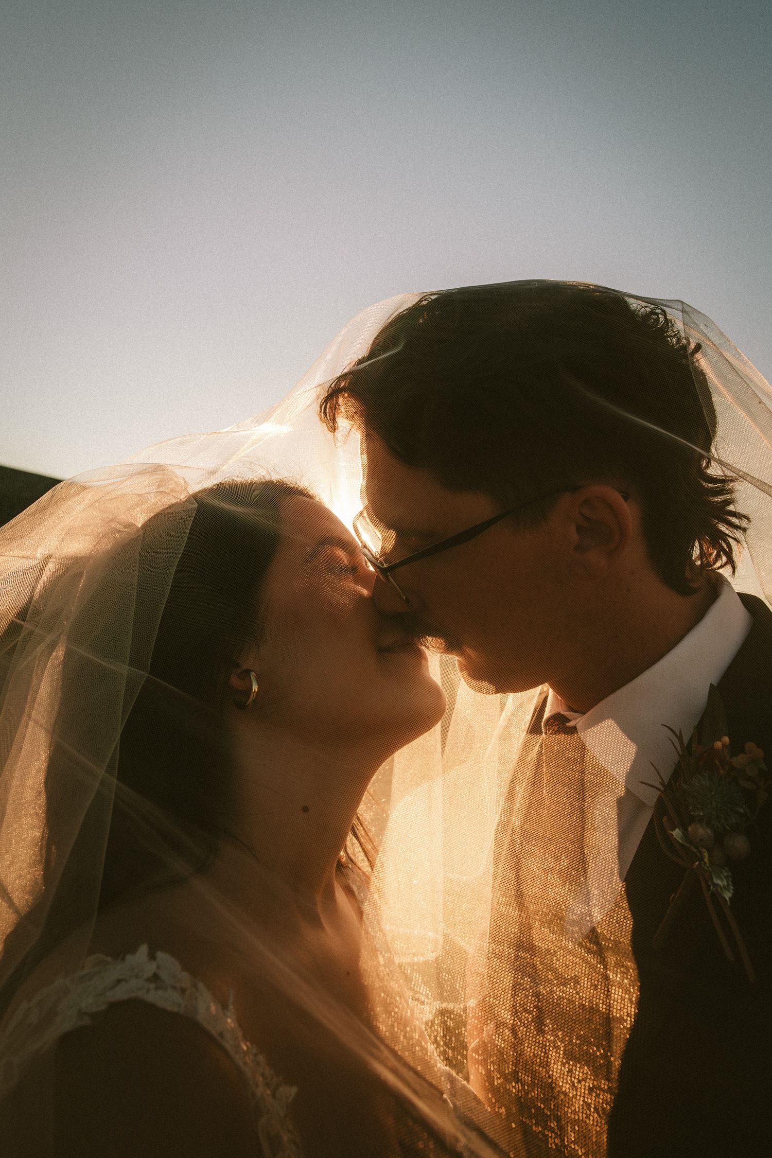 Romantic silhouette of bride and groom kissing under a veil with golden sunset backlight.