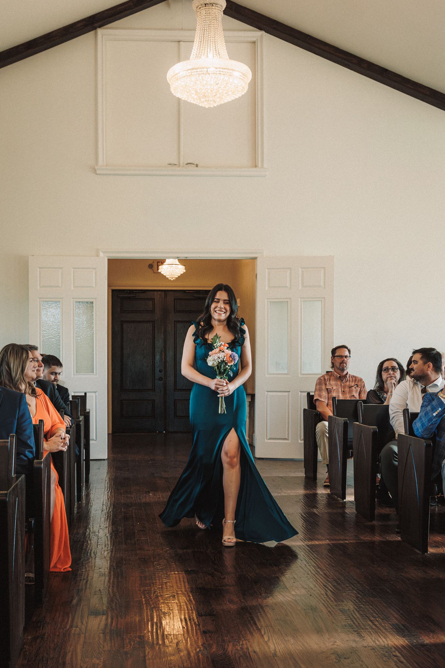 Smiling bridesmaid in teal gown walks down aisle of elegant white chapel with chandelier.