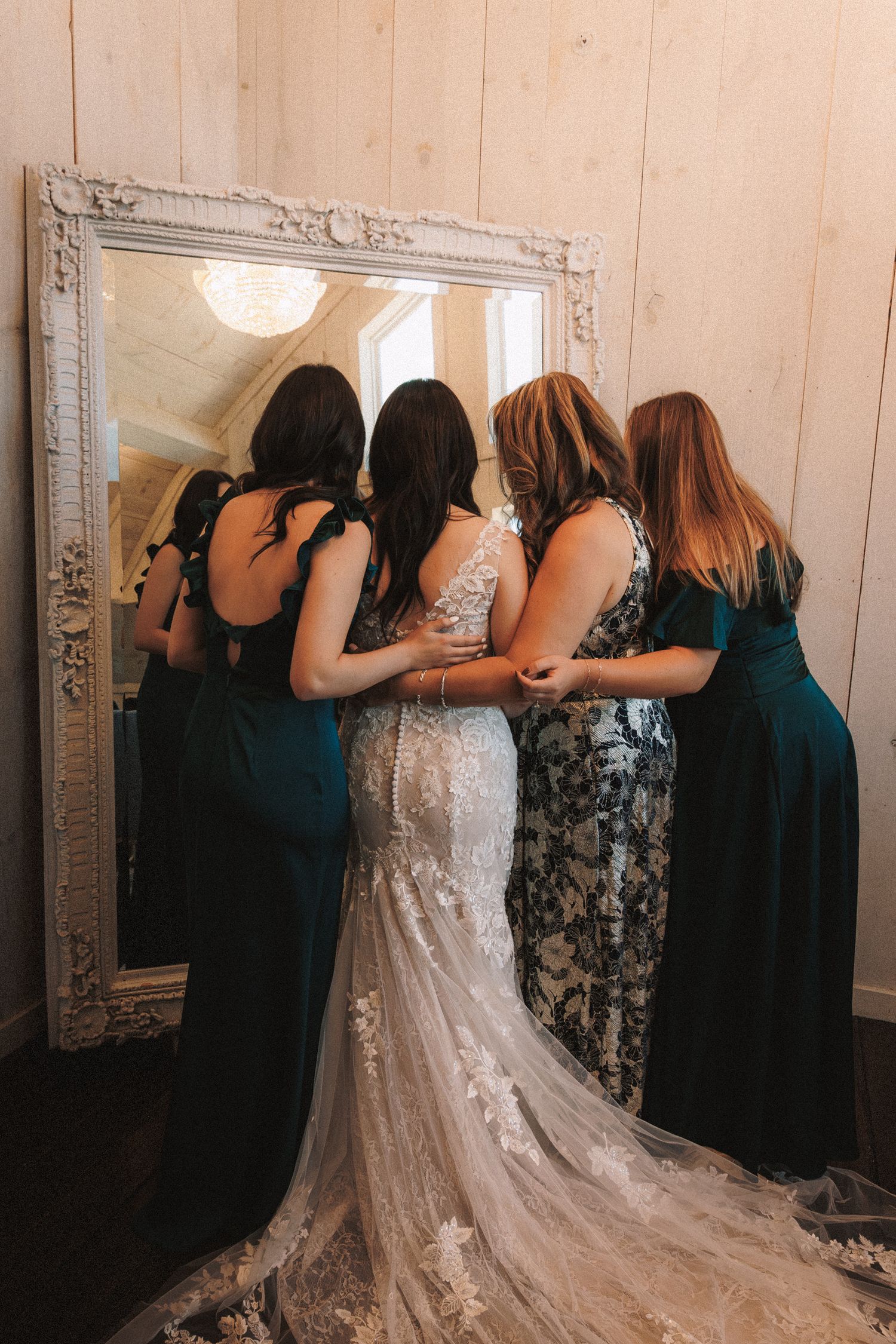 Bride in lace gown assisted by bridesmaids in emerald dresses while standing before an ornate mirror.