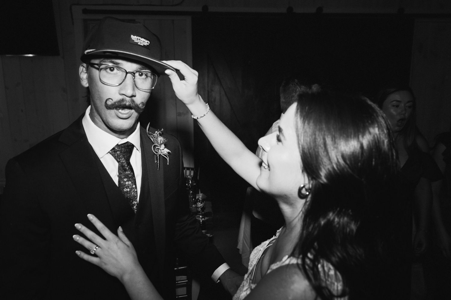 Black and white photo of bride adjusting grooms cap during wedding reception celebration.