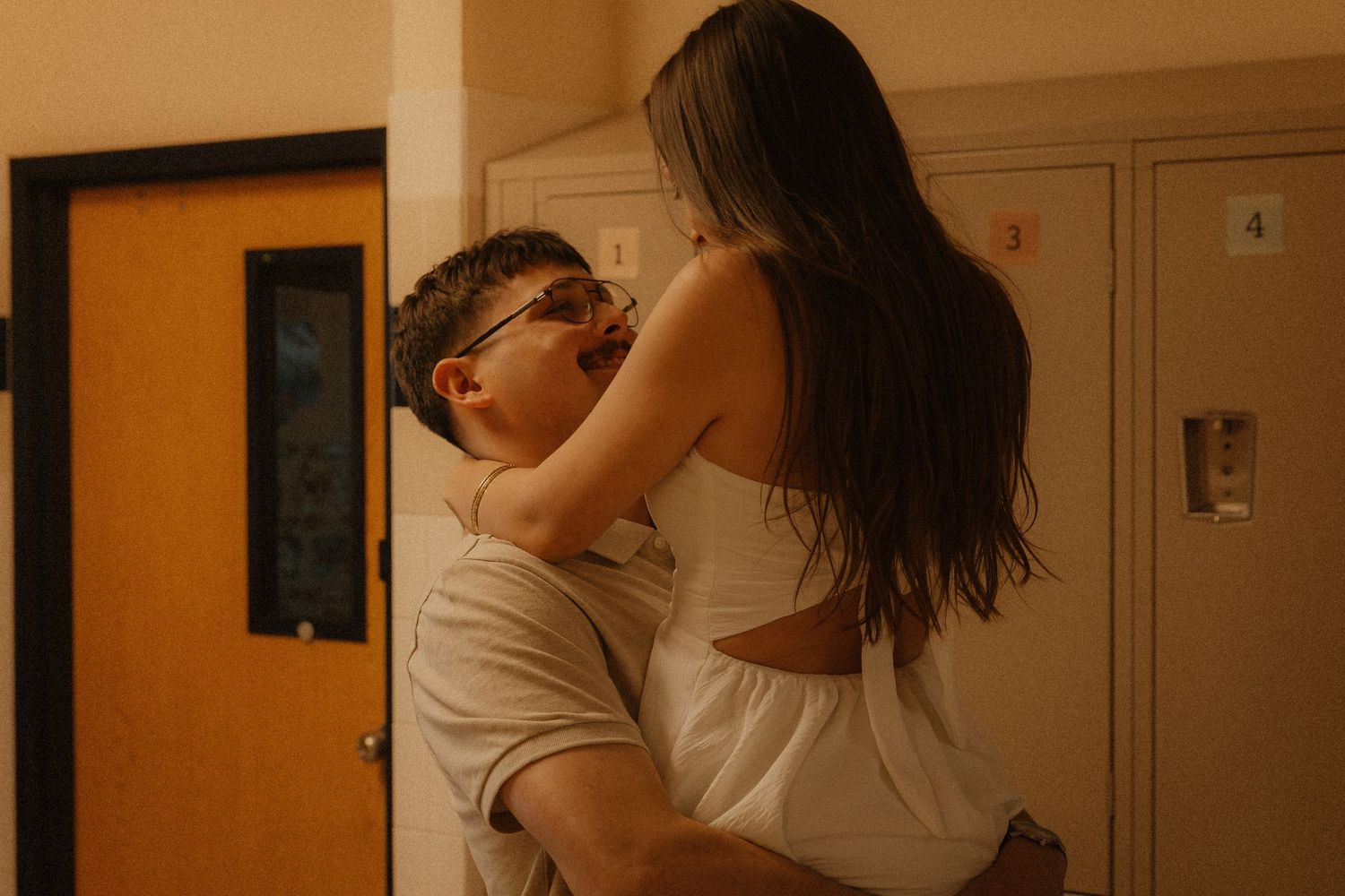 A couple sharing an intimate embrace near school lockers in a warmly lit hallway.
