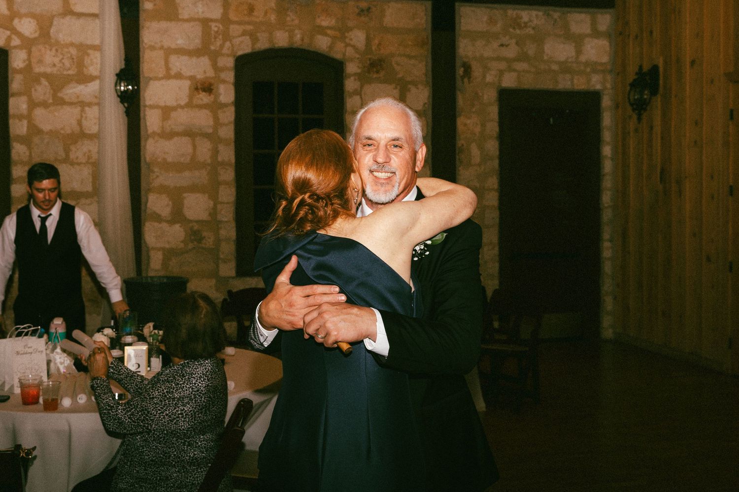 Father and bridesmaid share joyful dance embrace on stone-walled wedding reception dance floor.