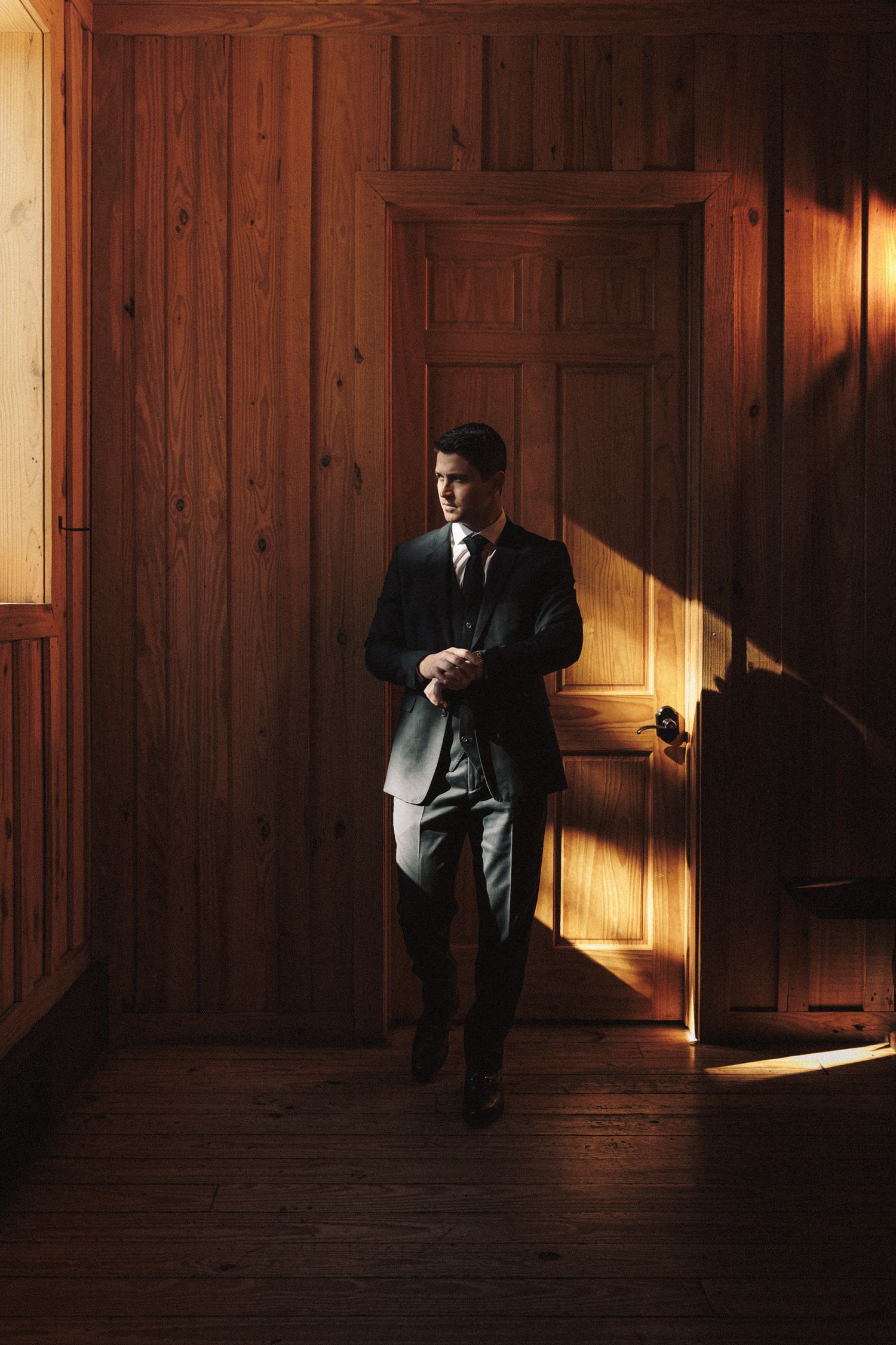 Dapper groom in dark suit leaning against wooden door in warm sunlit rustic interior setting.