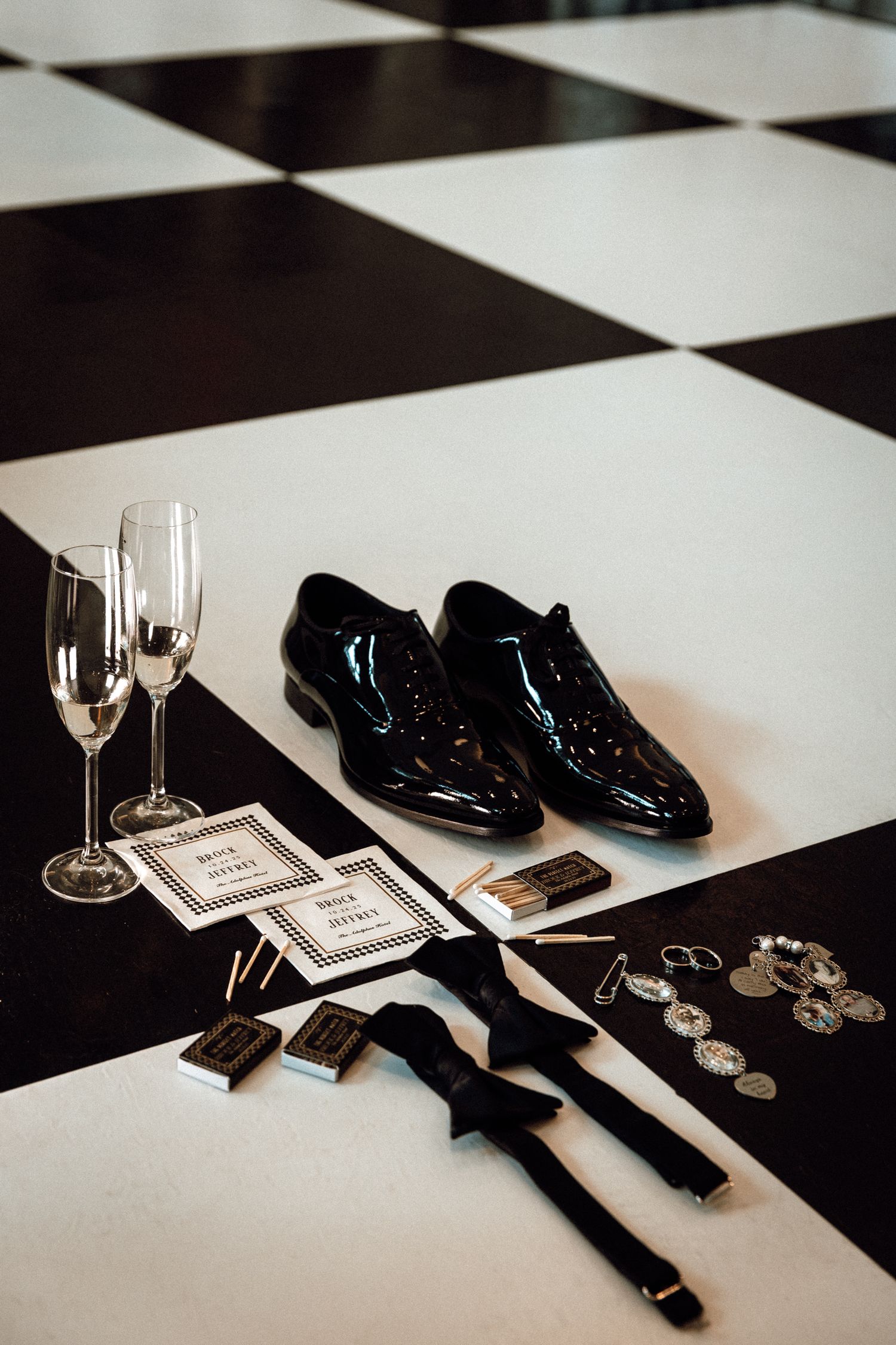 Black patent leather dress shoes with suspenders, cufflinks, and champagne flutes on a checkered floor.