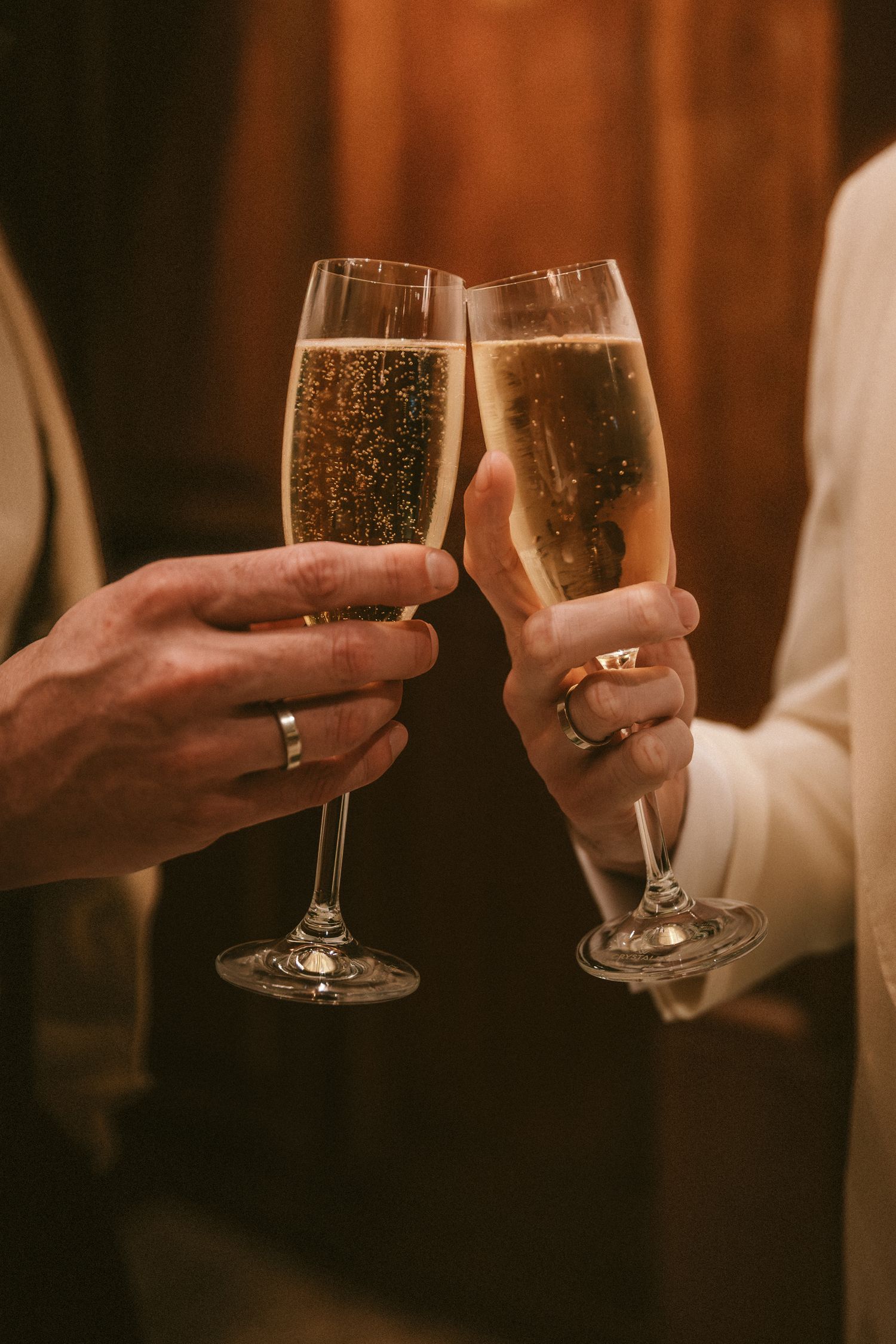 Newlywed couple clinking champagne flutes in celebration, both wearing wedding bands.