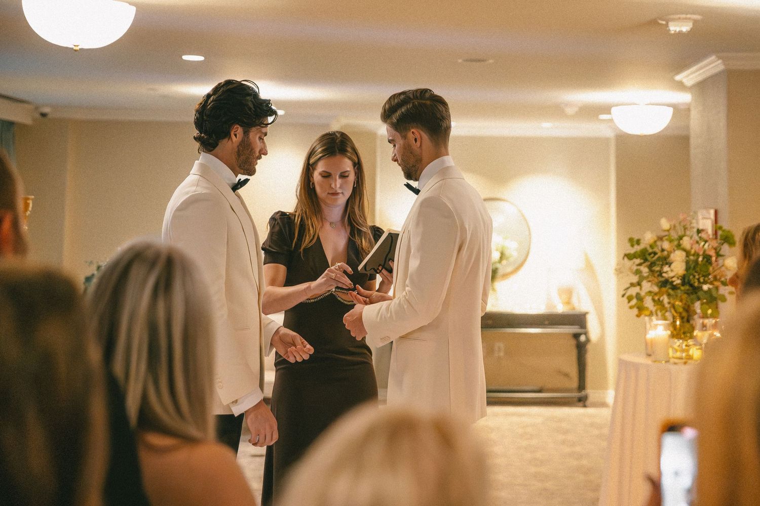 Two grooms exchanging vows with a female officiant during their indoor wedding ceremony.