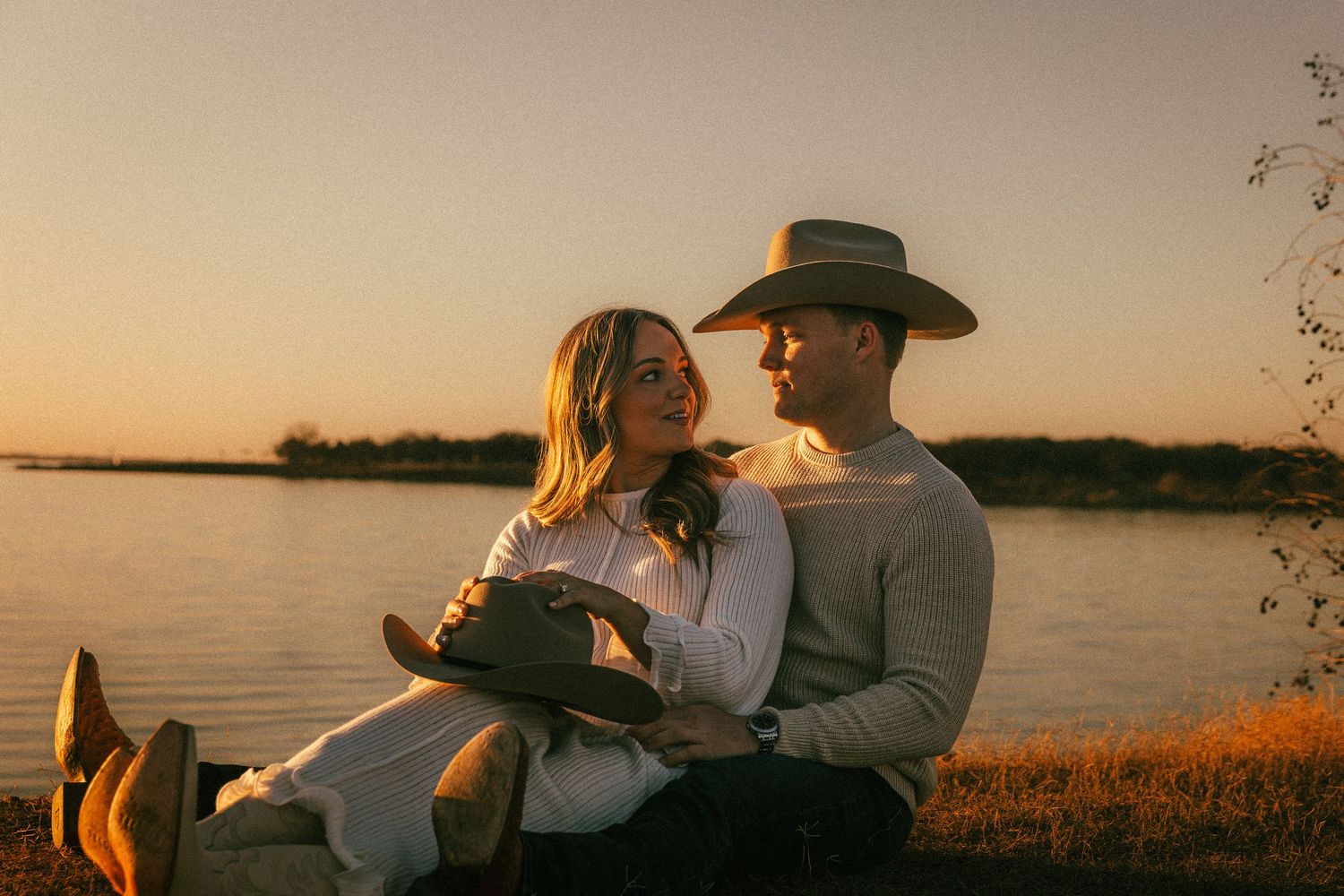 Romantic couple sitting by a lake at sunset with a cowboy hat silhouetted against the golden sky.
