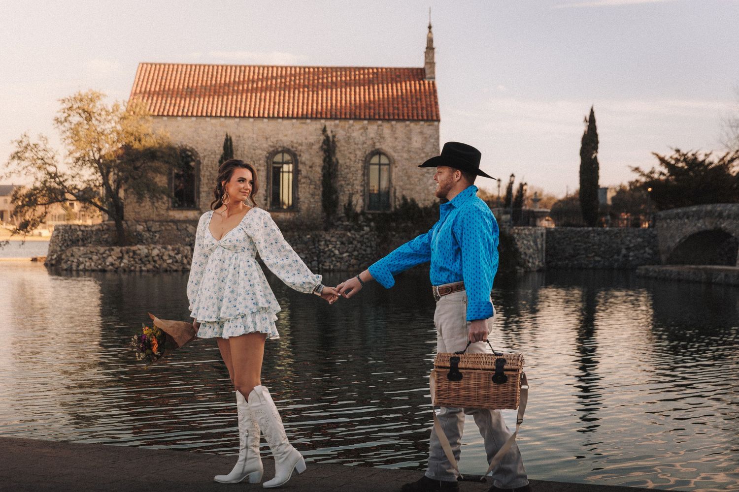A couple in western-inspired outfits dances by a waterfront at sunset with a historic stone church in the background.