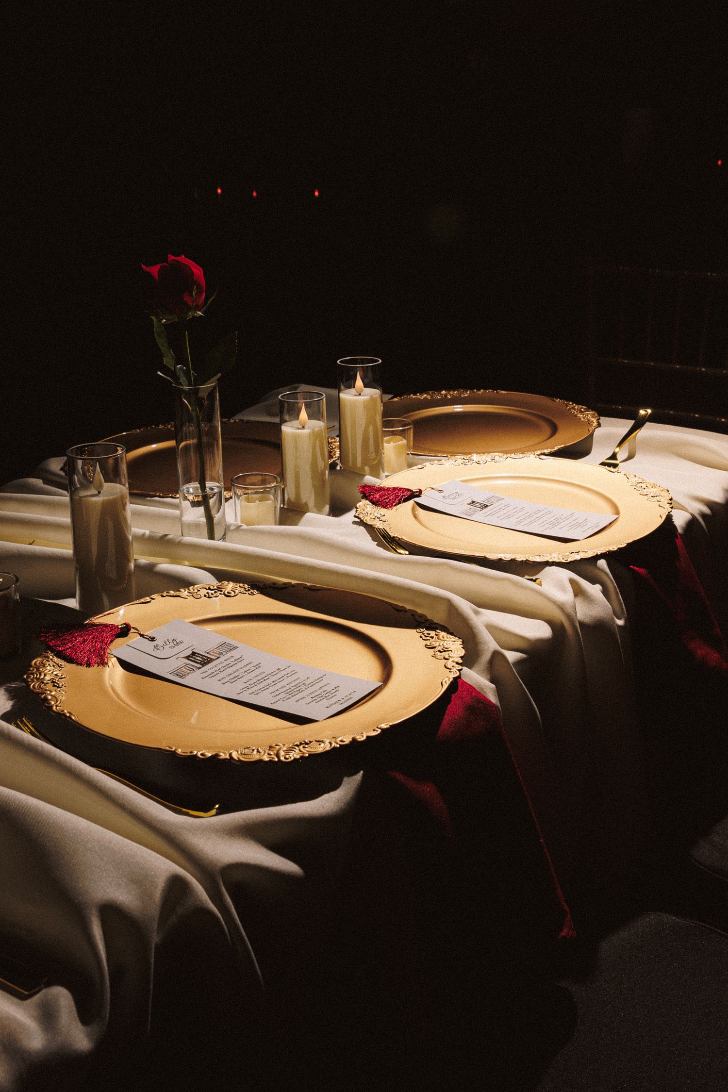 Elegant wedding reception table with gold charger plates, candles, red roses, and white linen in dim lighting.