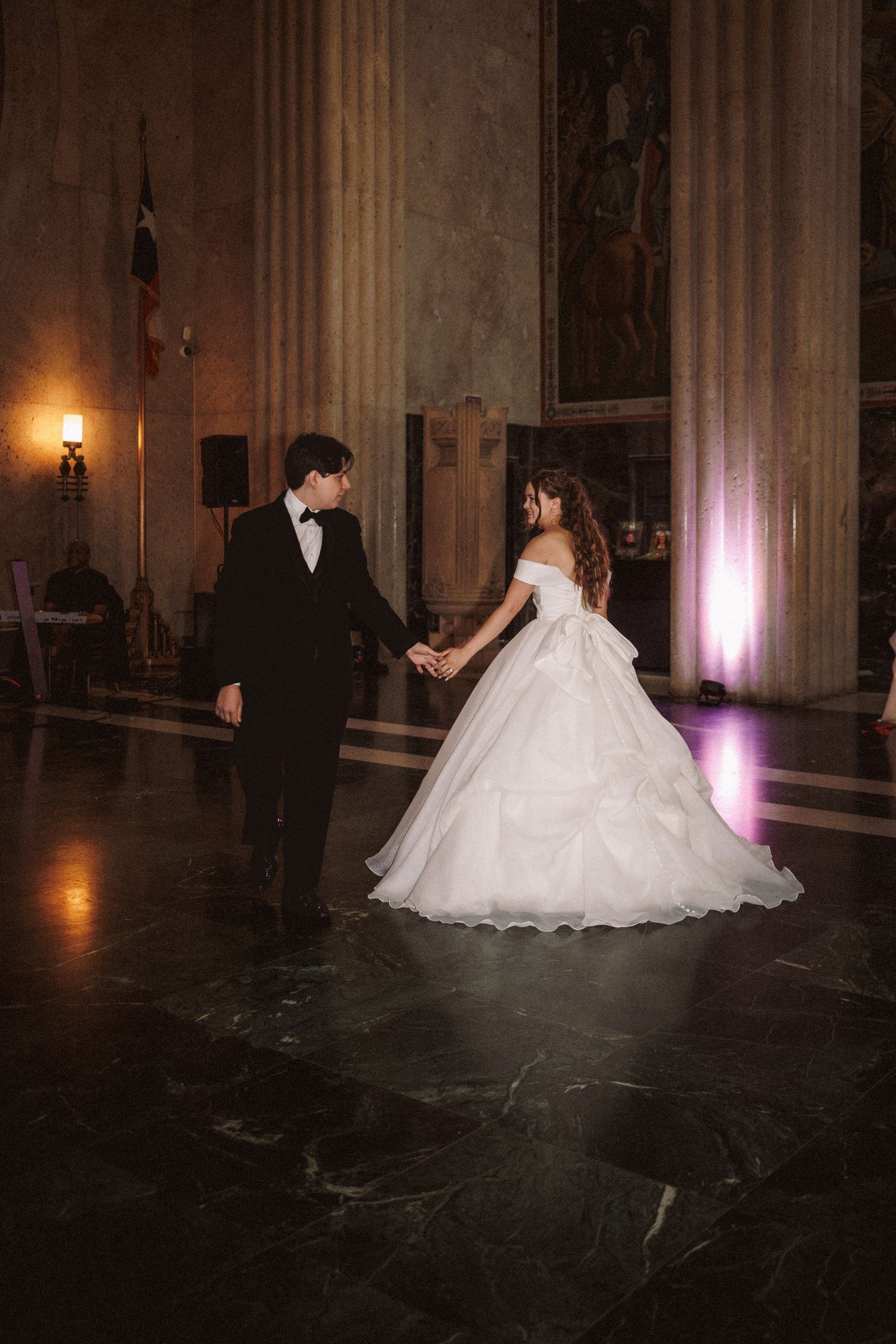 Bride and groom hold hands during first dance in grand marble hall with ornate columns.