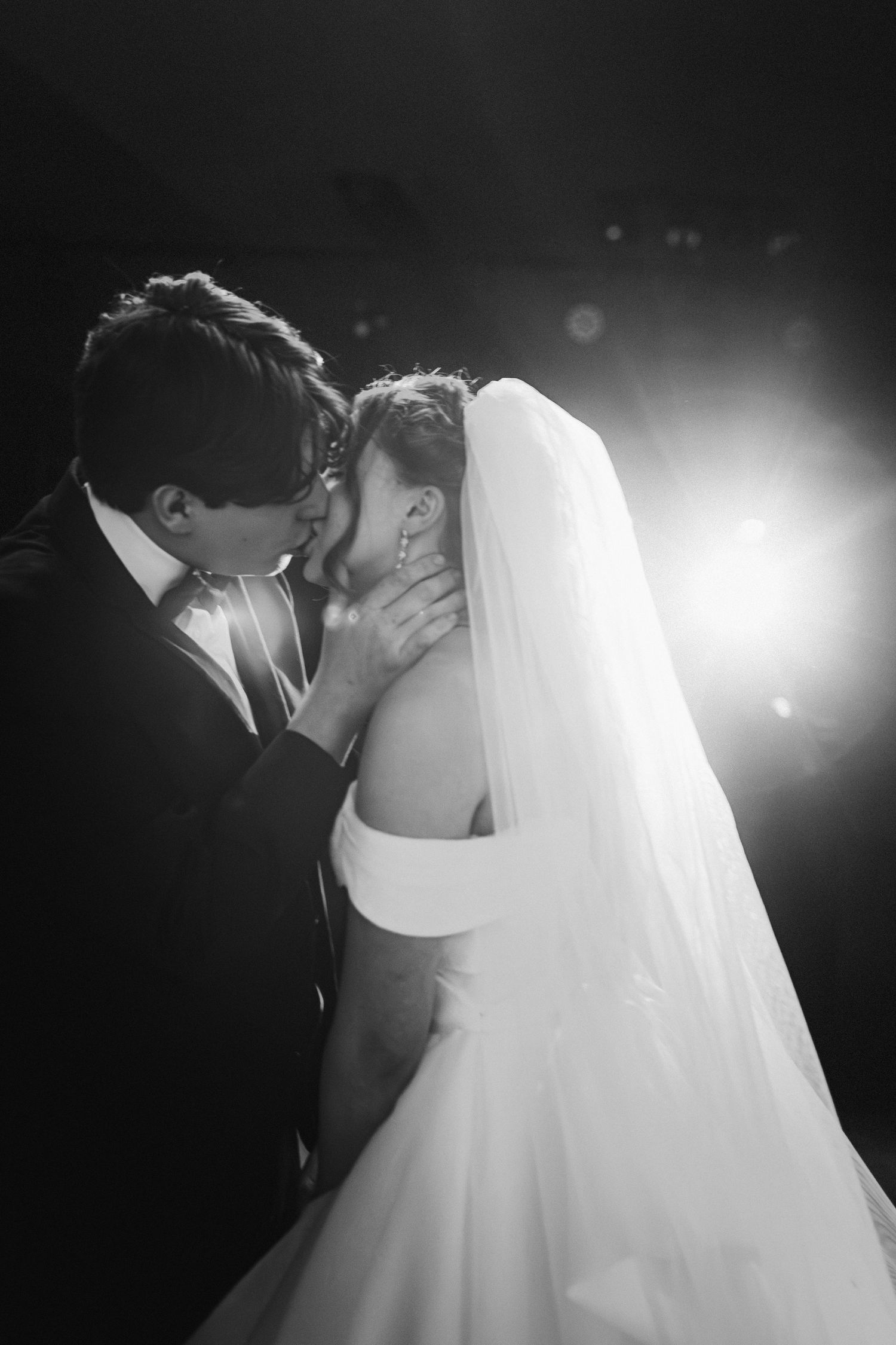 Black and white photo of bride and groom sharing romantic kiss with dramatic backlight.