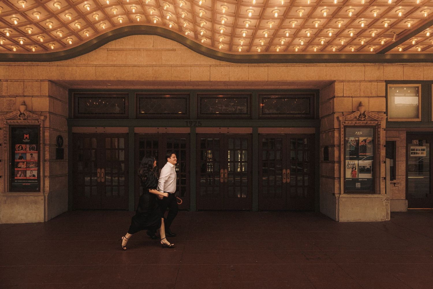 Couple walking past illuminated theater entrance with ornate ceiling and grand doors at night.