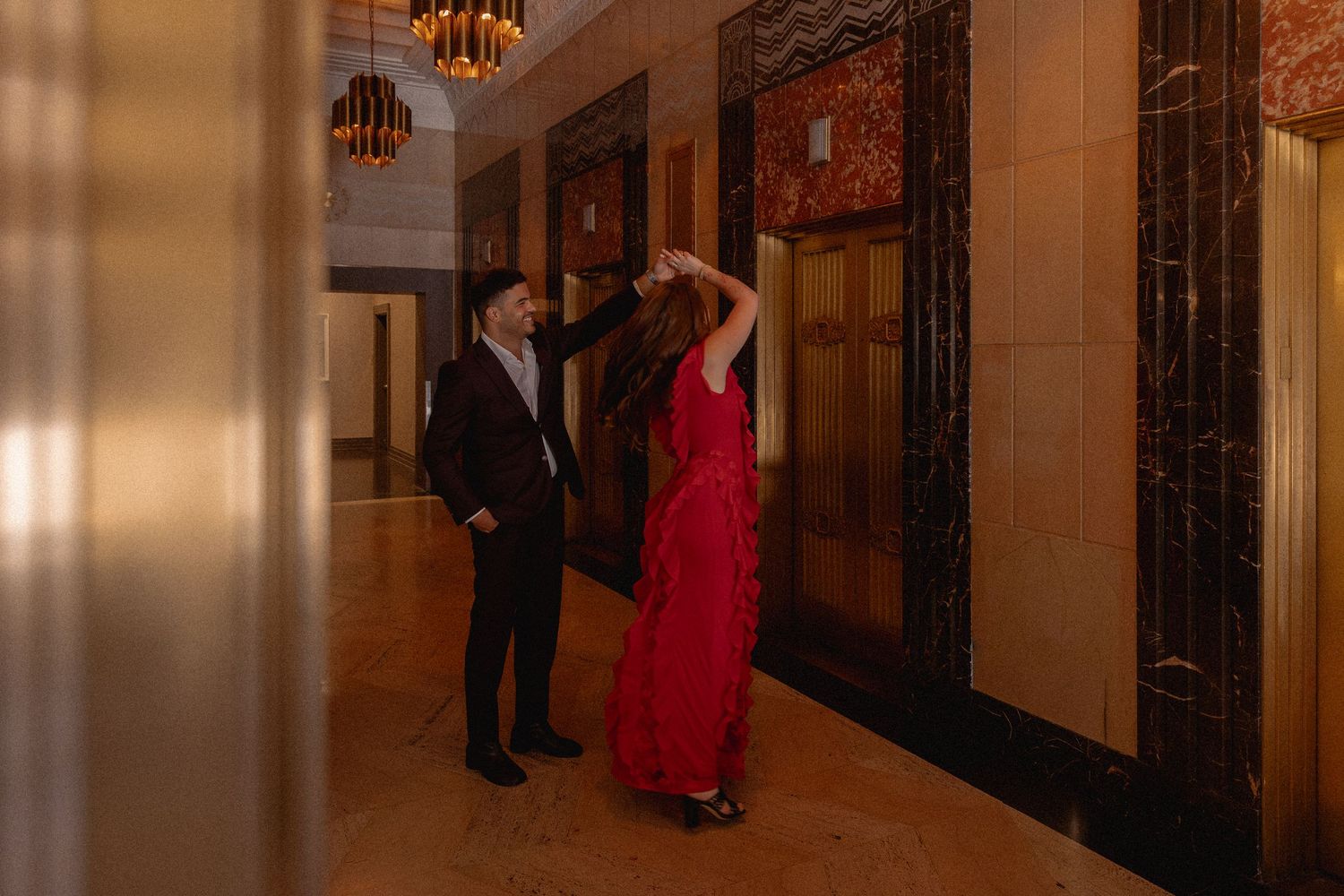 Couple in elegant attire dancing in a grand hallway with marble walls and chandeliers.