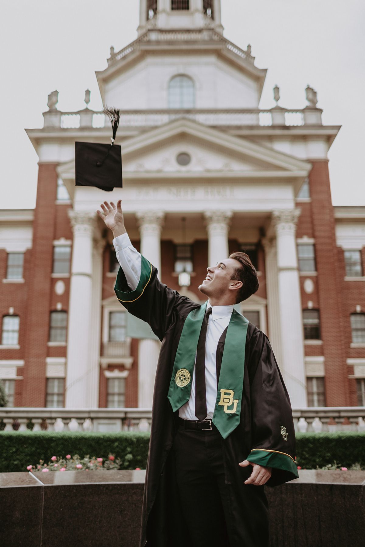 College graduate in green and black regalia celebrates by tossing graduation cap in front of classical campus building.