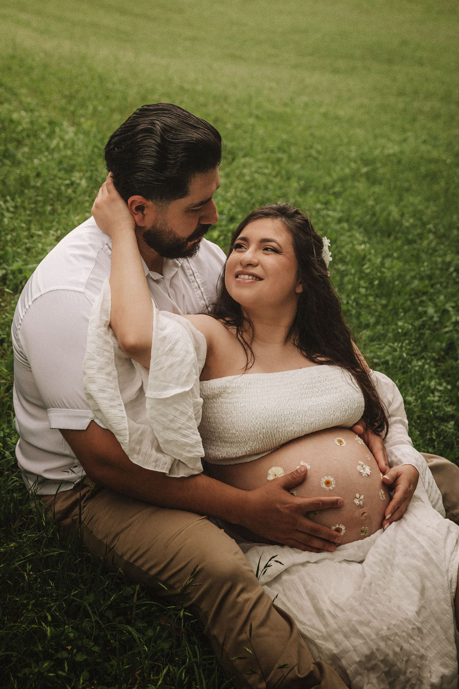 Intimate maternity photo sequence shows a couple in white embracing on green grass during golden hour.