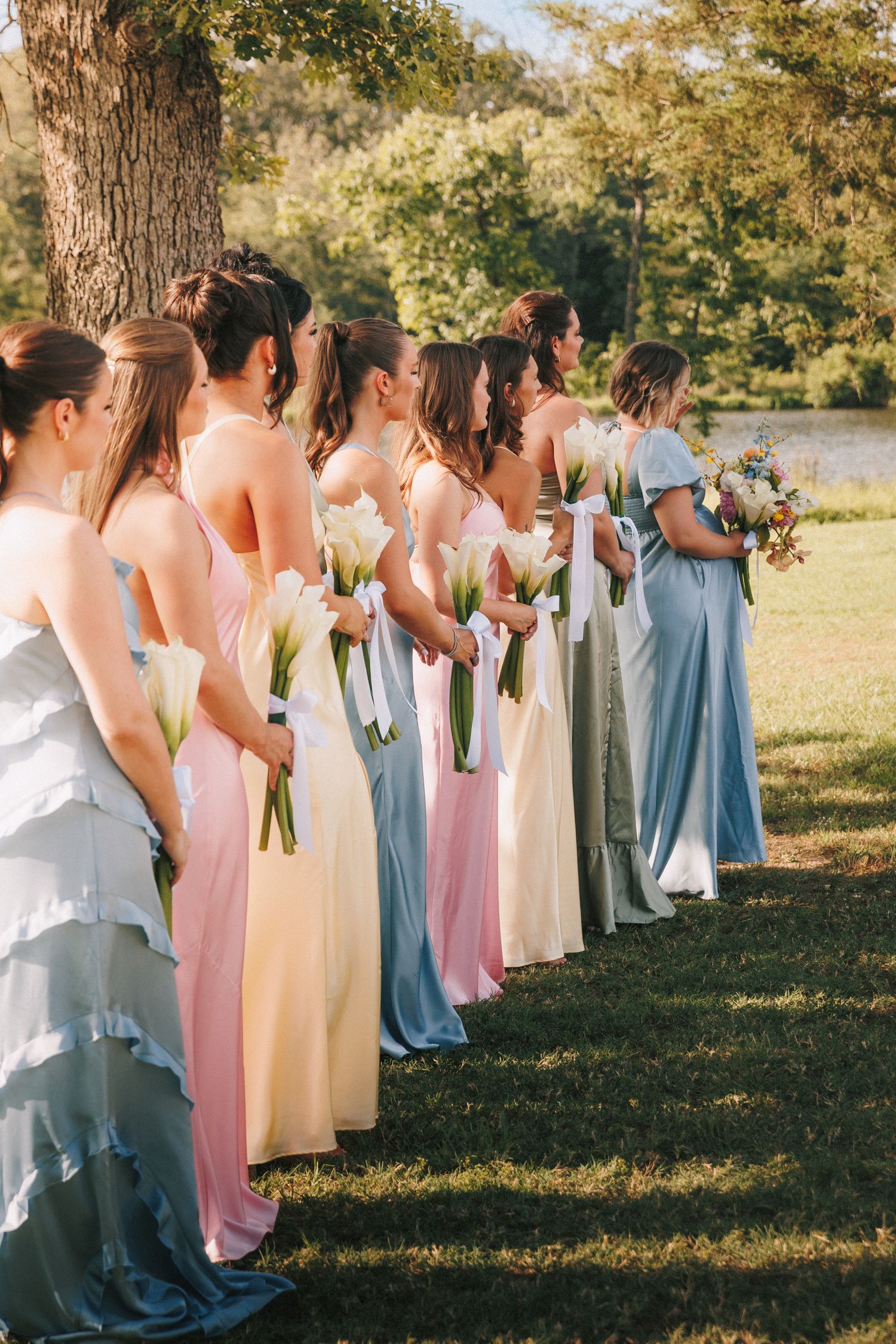 Bridesmaids wearing pastel blue and pink dresses stand in a line holding bouquets on a sunny green lawn by a lake.