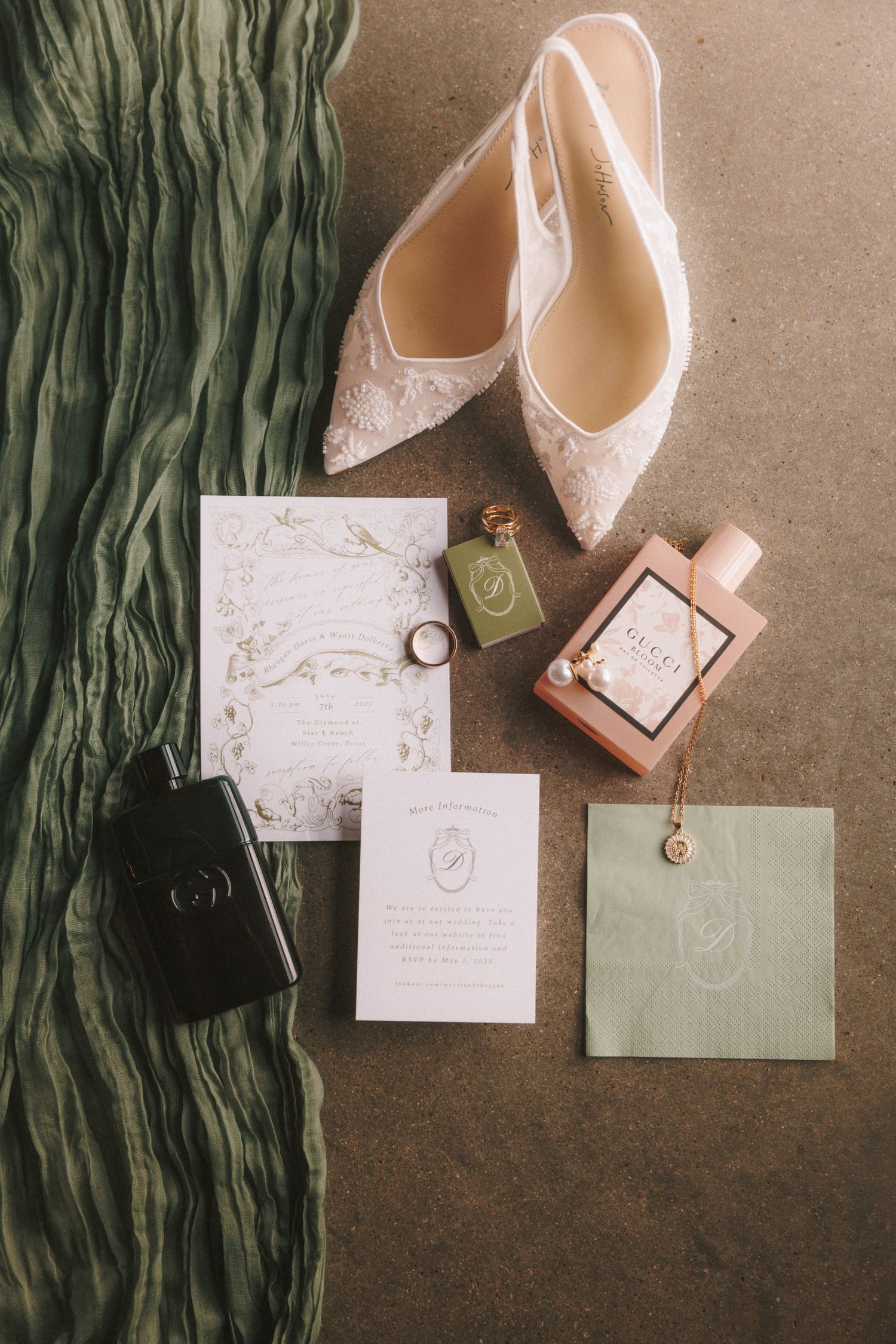 Wedding accessories including white lace heels, invitations, and a perfume bottle arranged on a dark surface with green fabric.
