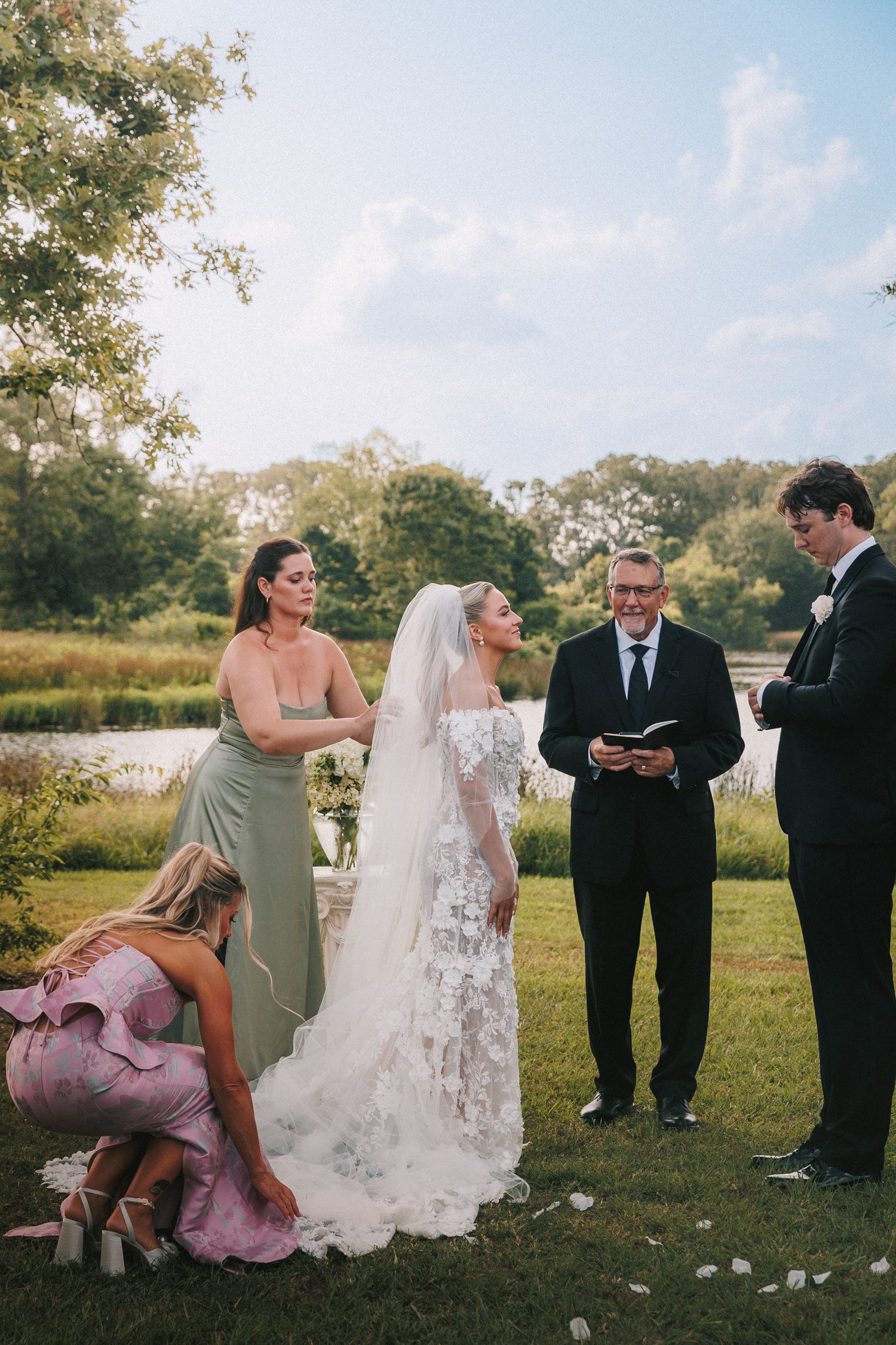 A wedding ceremony takes place outdoors on a sunny day with guests and officiant.