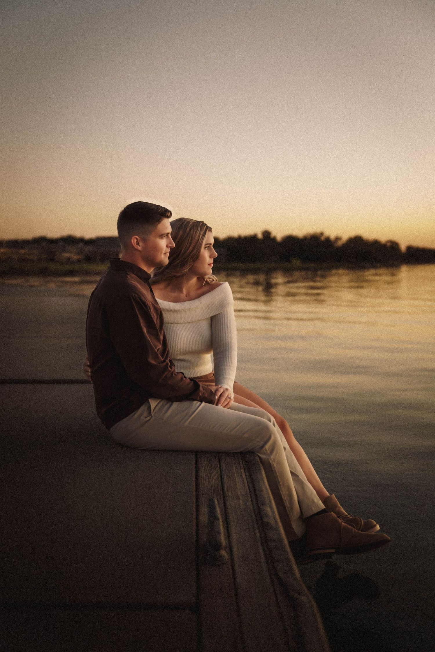 A couple shares an intimate moment sitting on a dock at sunset overlooking a tranquil lake.