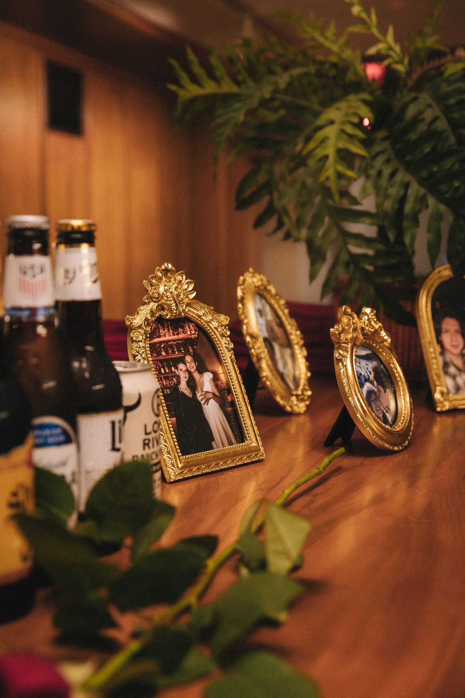 Gold ornate picture frames with photos displayed on a wooden table beside beer bottles and roses at a wedding reception.