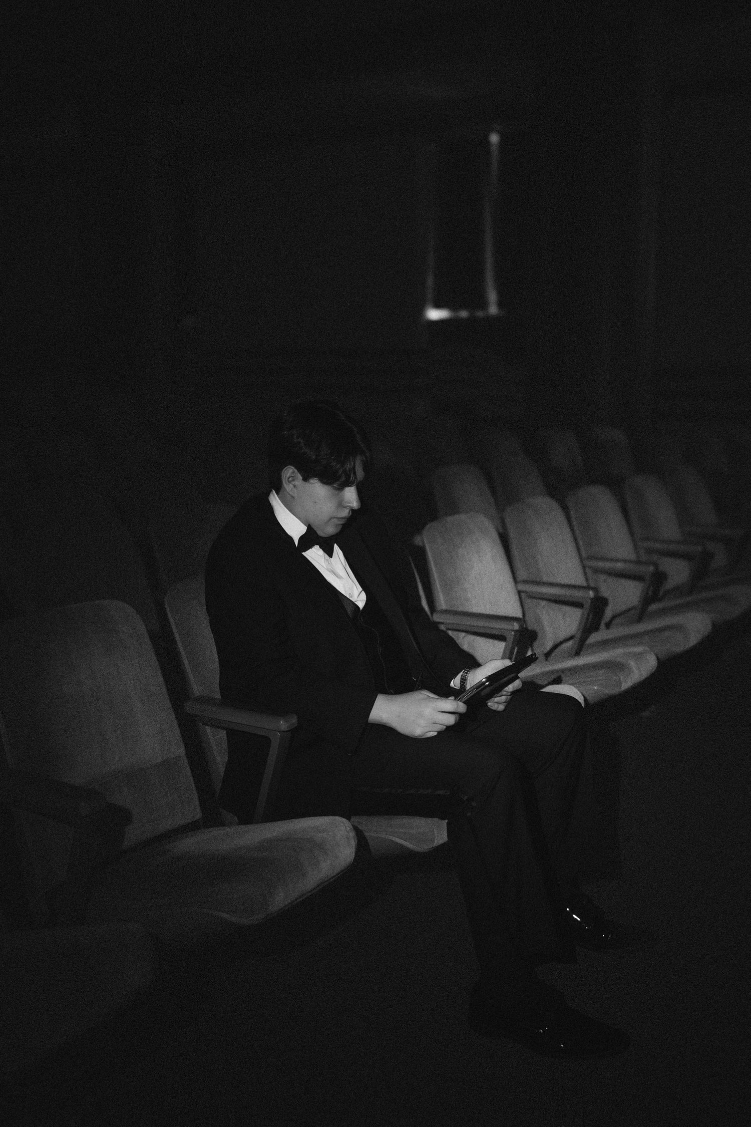 Groom in black tuxedo reading alone in dark empty theater seats, black and white.