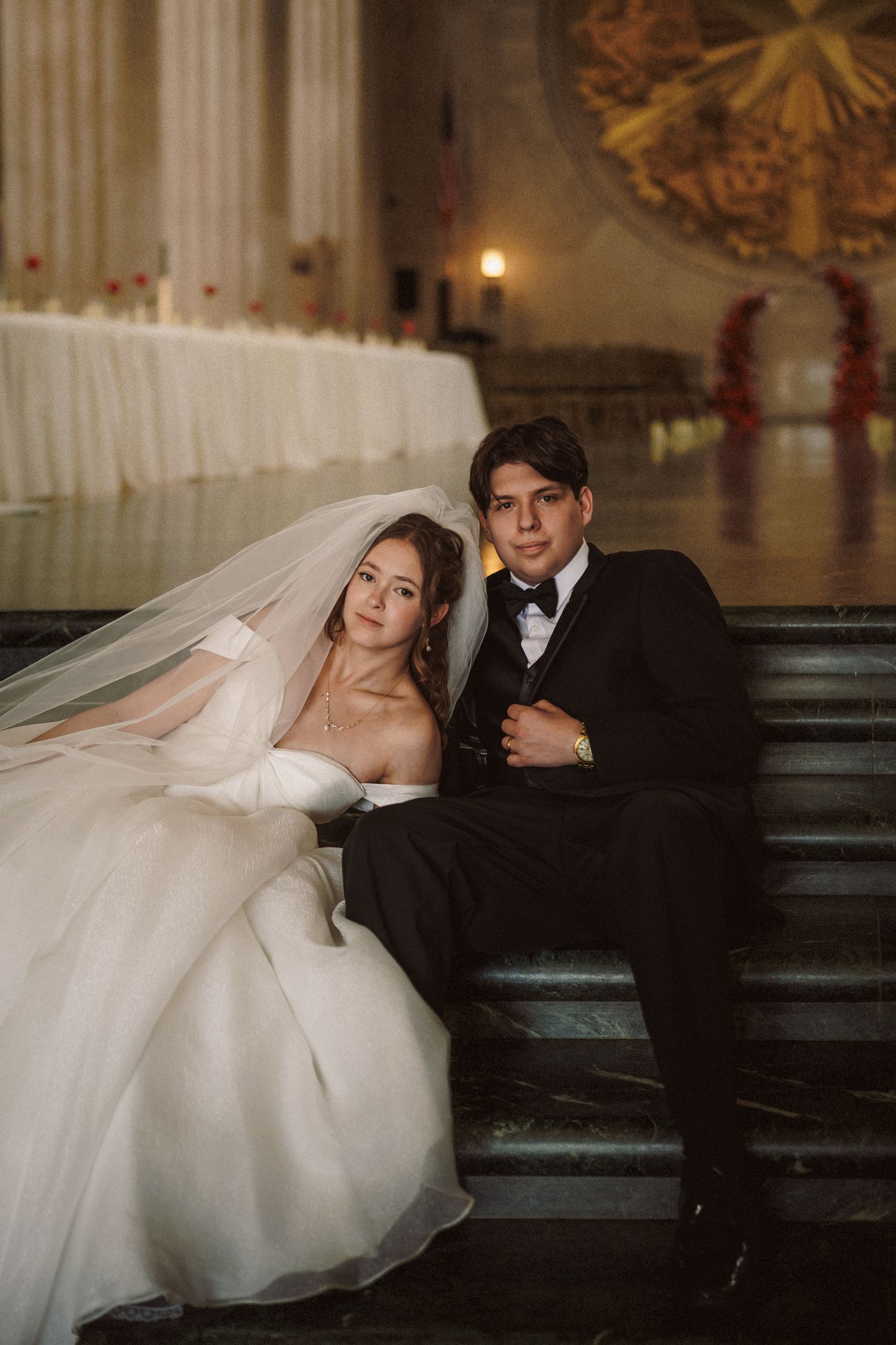 Bride in white ballgown and veil sits with groom in black tuxedo on marble steps in grand wedding venue.
