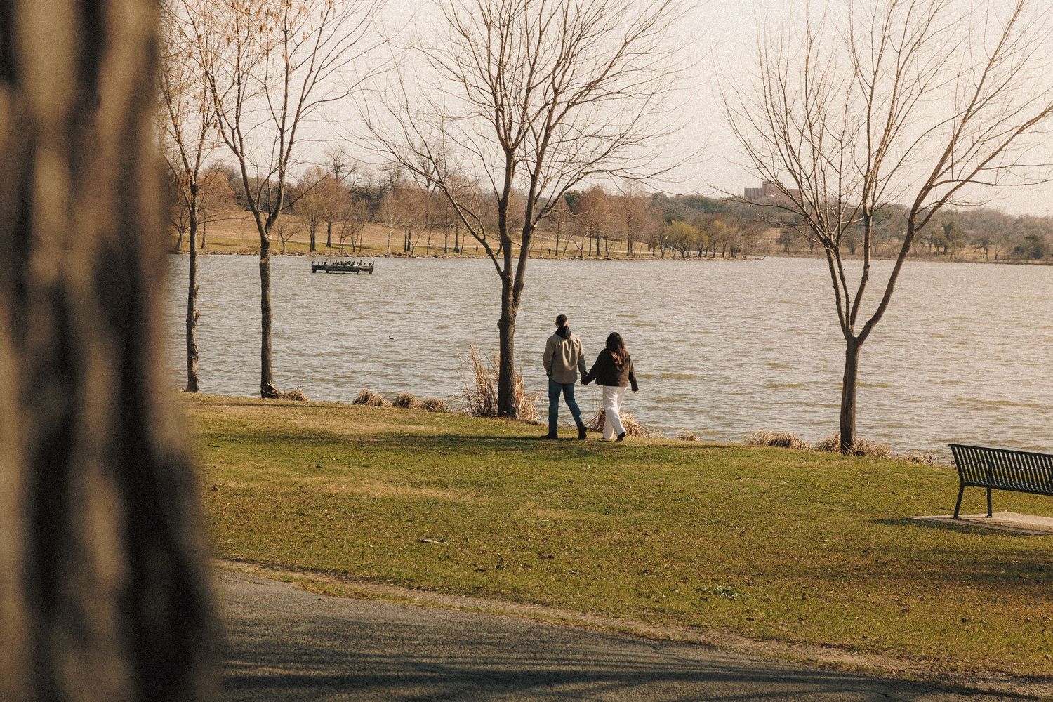 A couple strolls along the edge of a serene lake on a winter day with bare trees and grass in the foreground.