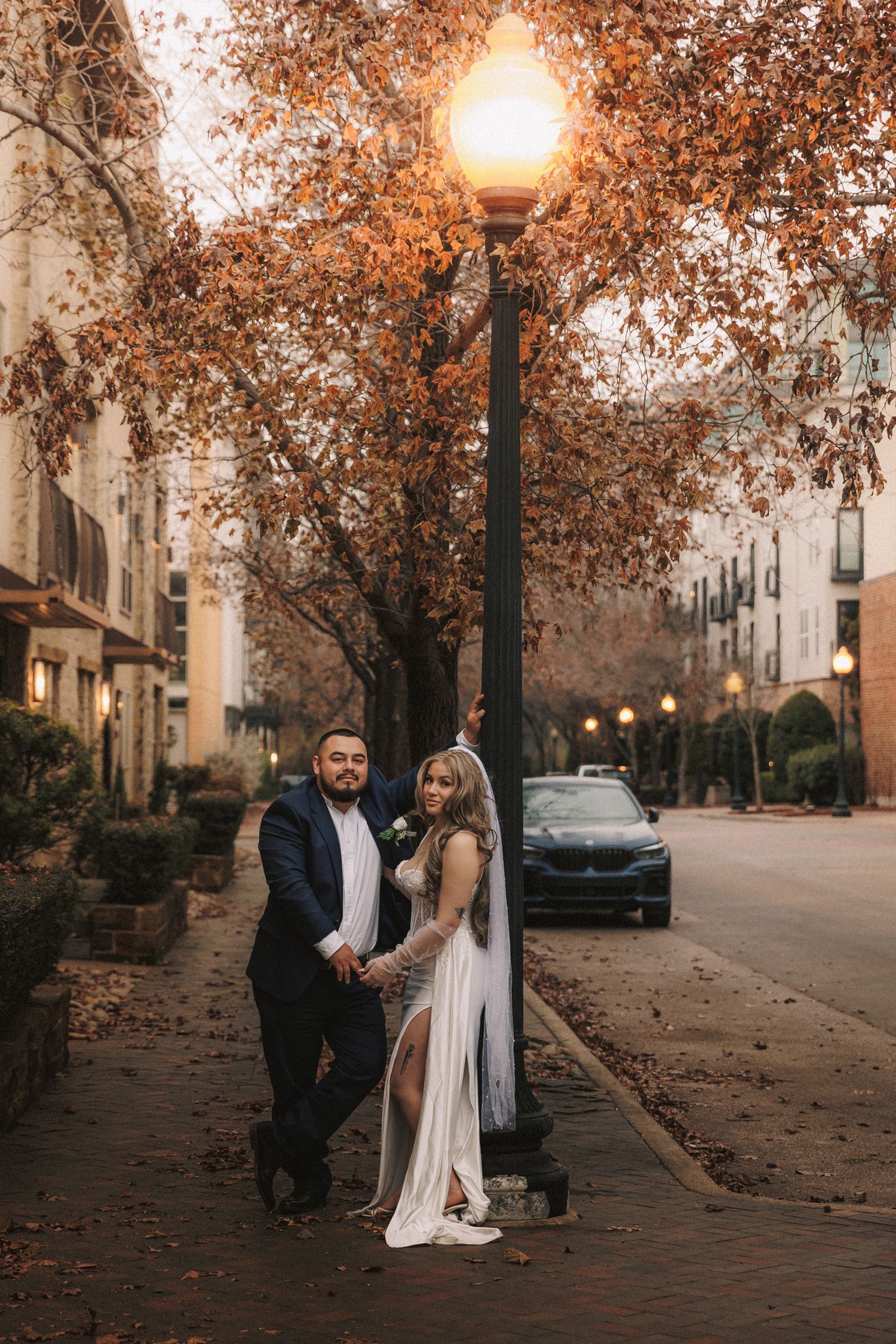 A couple shares a romantic moment on a tree-lined street during autumn at dusk.