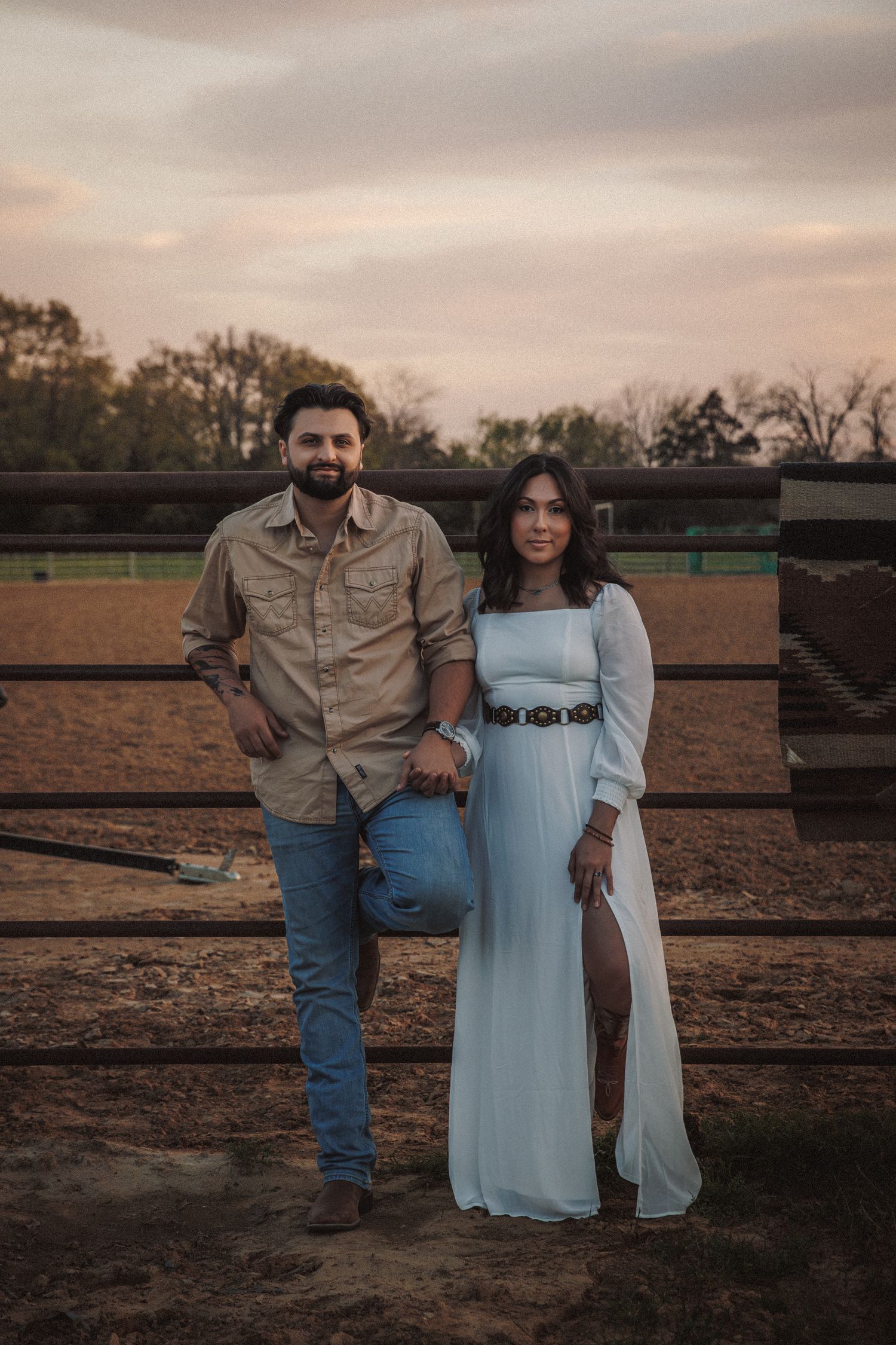 Stylish couple posing at sunset by rustic ranch fence, woman in white dress with belt.