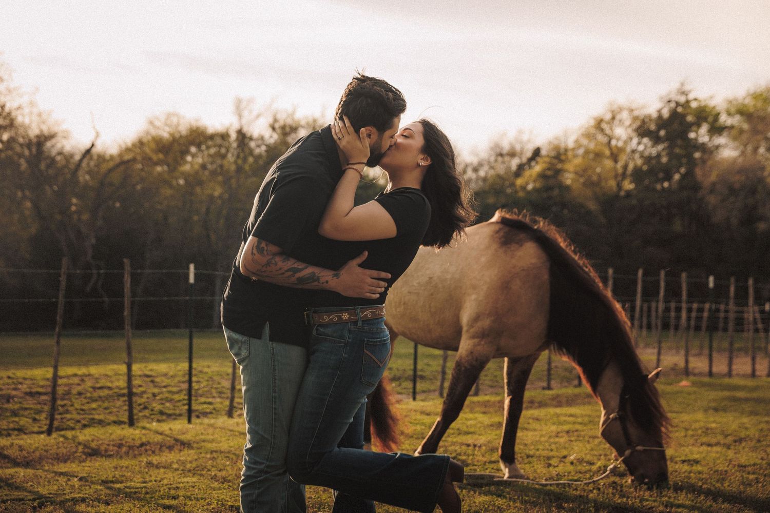 Couple sharing a romantic kiss in a sunlit farm field with a brown horse grazing beside them.