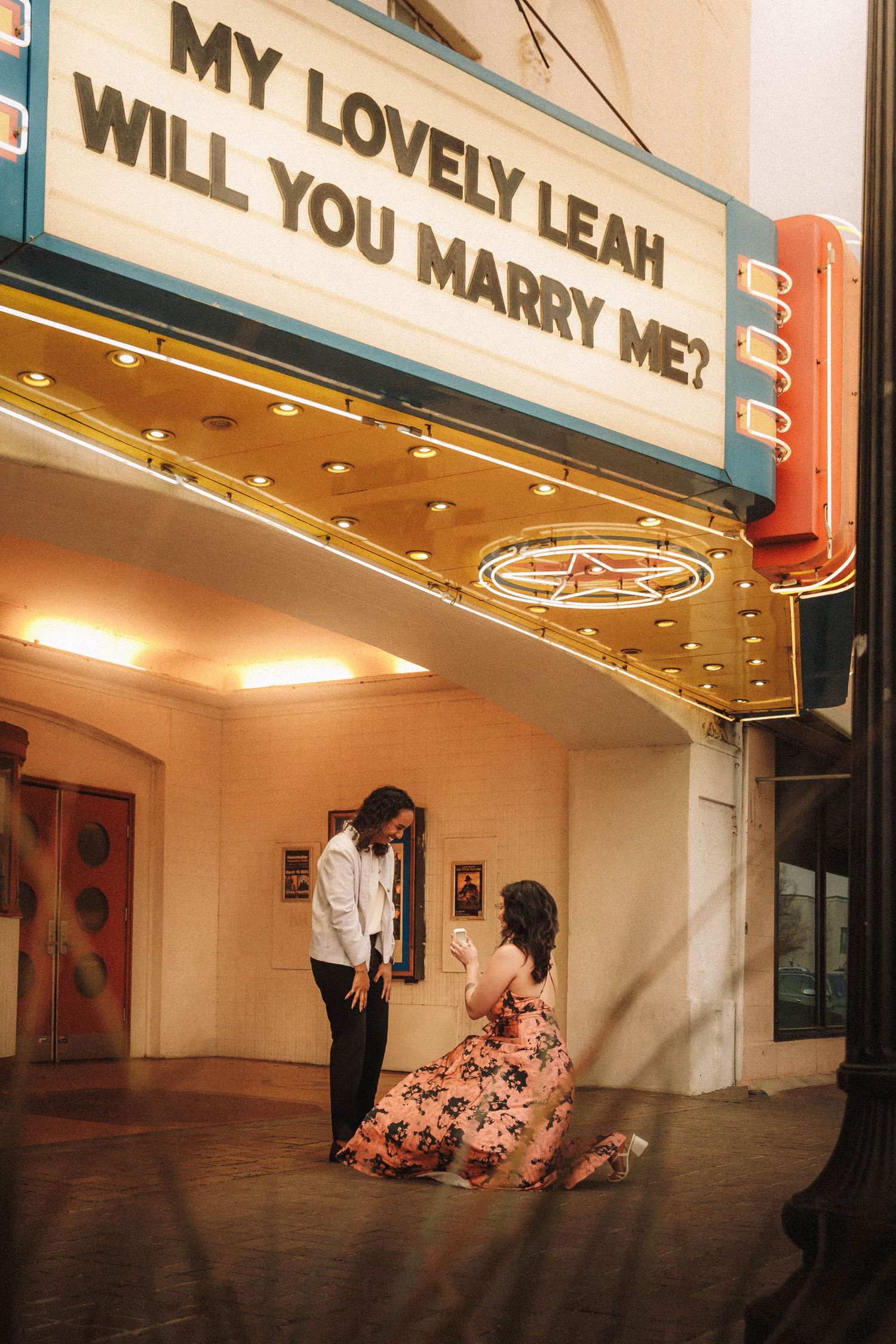 Woman in floral dress on knees during marriage proposal under lit theater marquee reading My Lovely Leah Will You Marry Me.