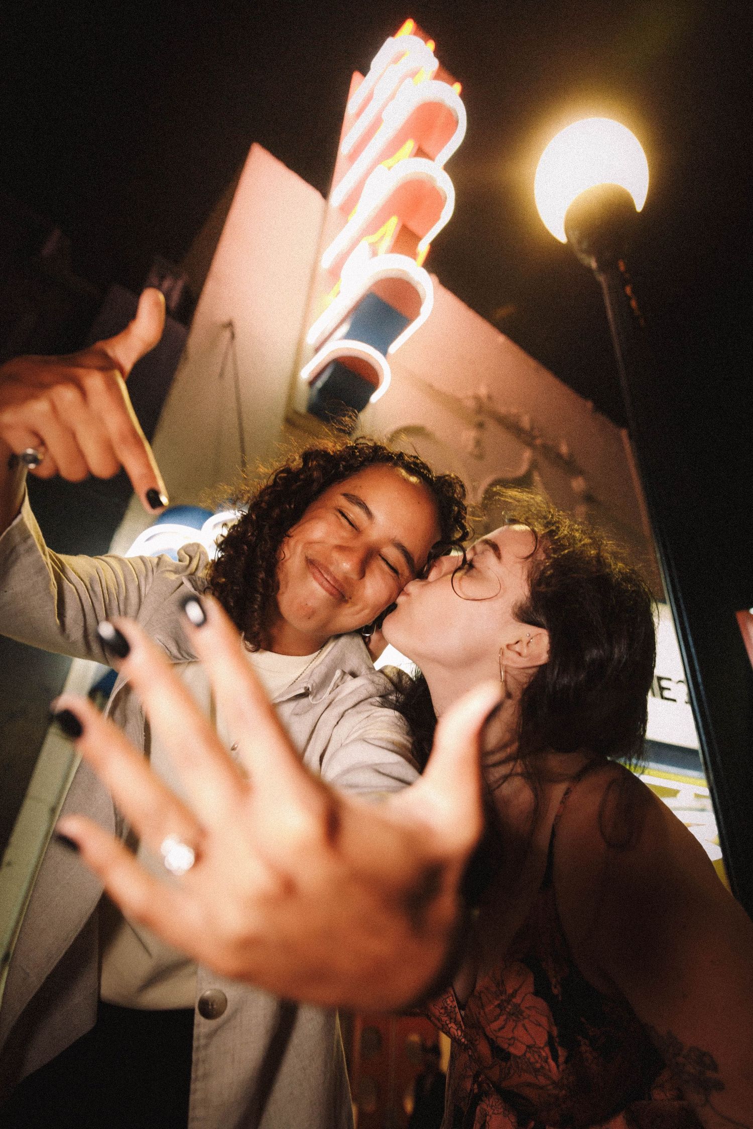 Happy couple takes a selfie with a kiss outside a neon-lit venue at night.